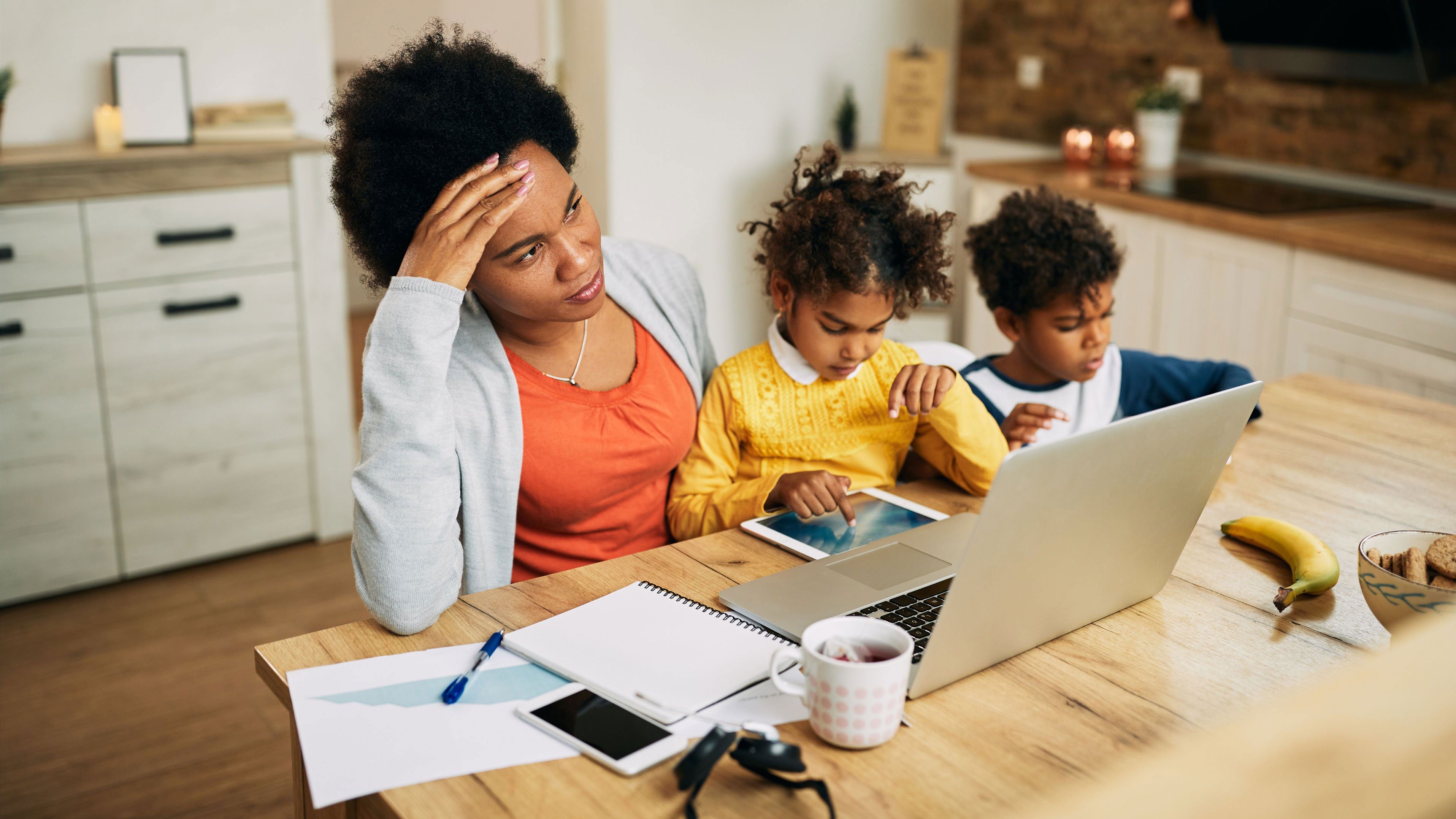 A mother works on her laptop while entertaining her two children at the kitchen table.