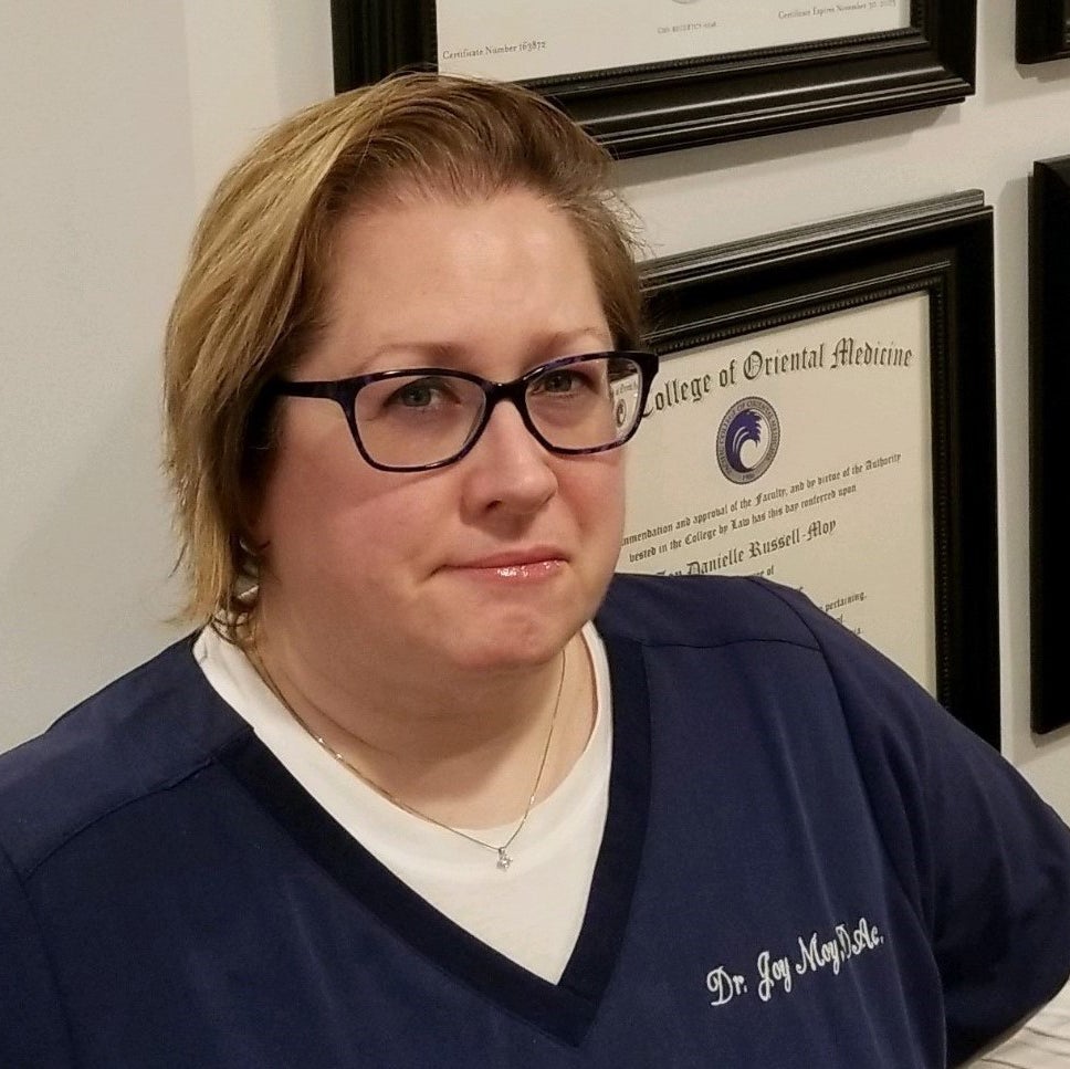 Doctor Joy Moy poses for a picture wearing black-framed glasses and blue scrubs.