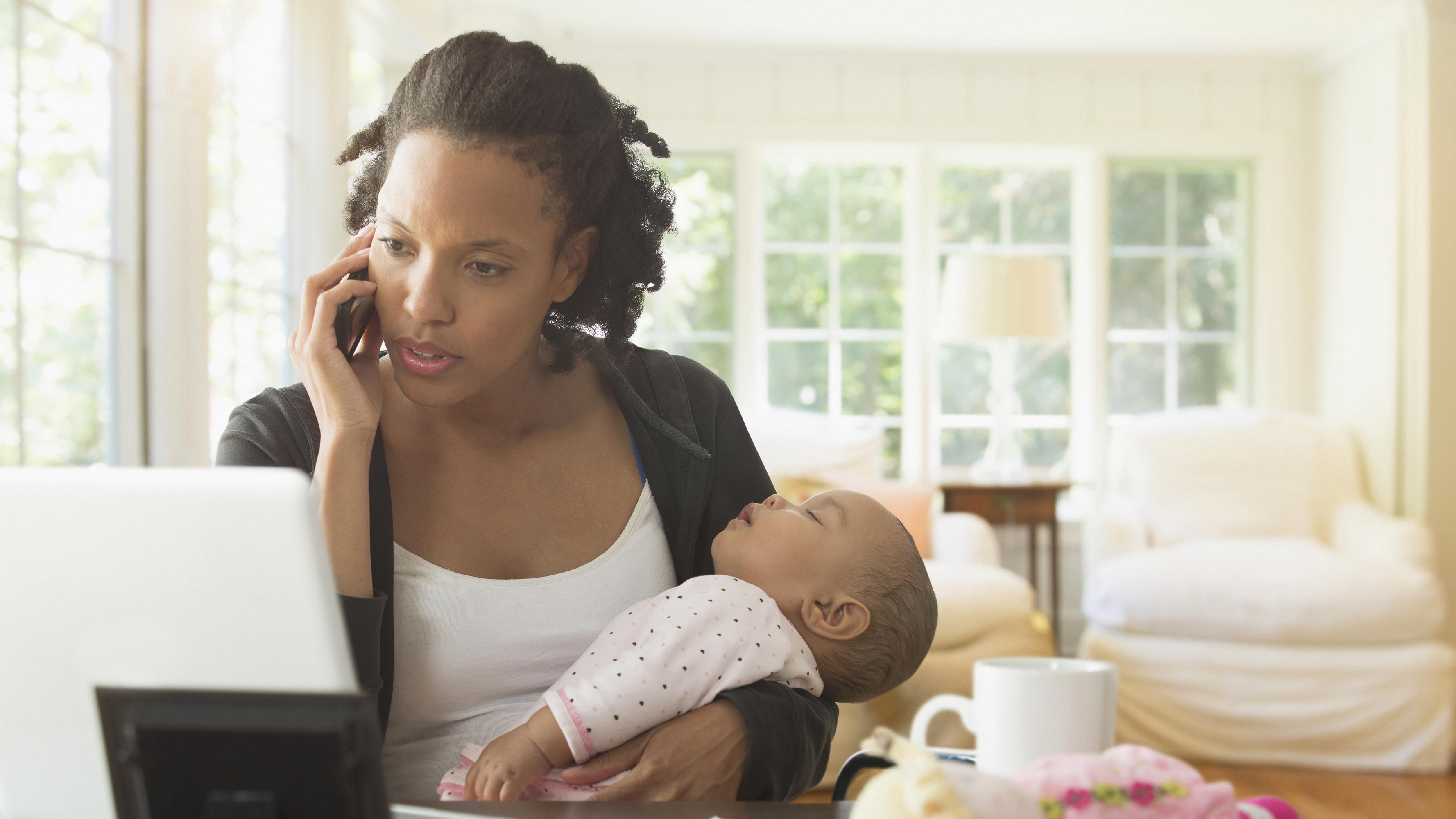 A mother uses her laptop and holds her sleeping infant while talking on the phone.