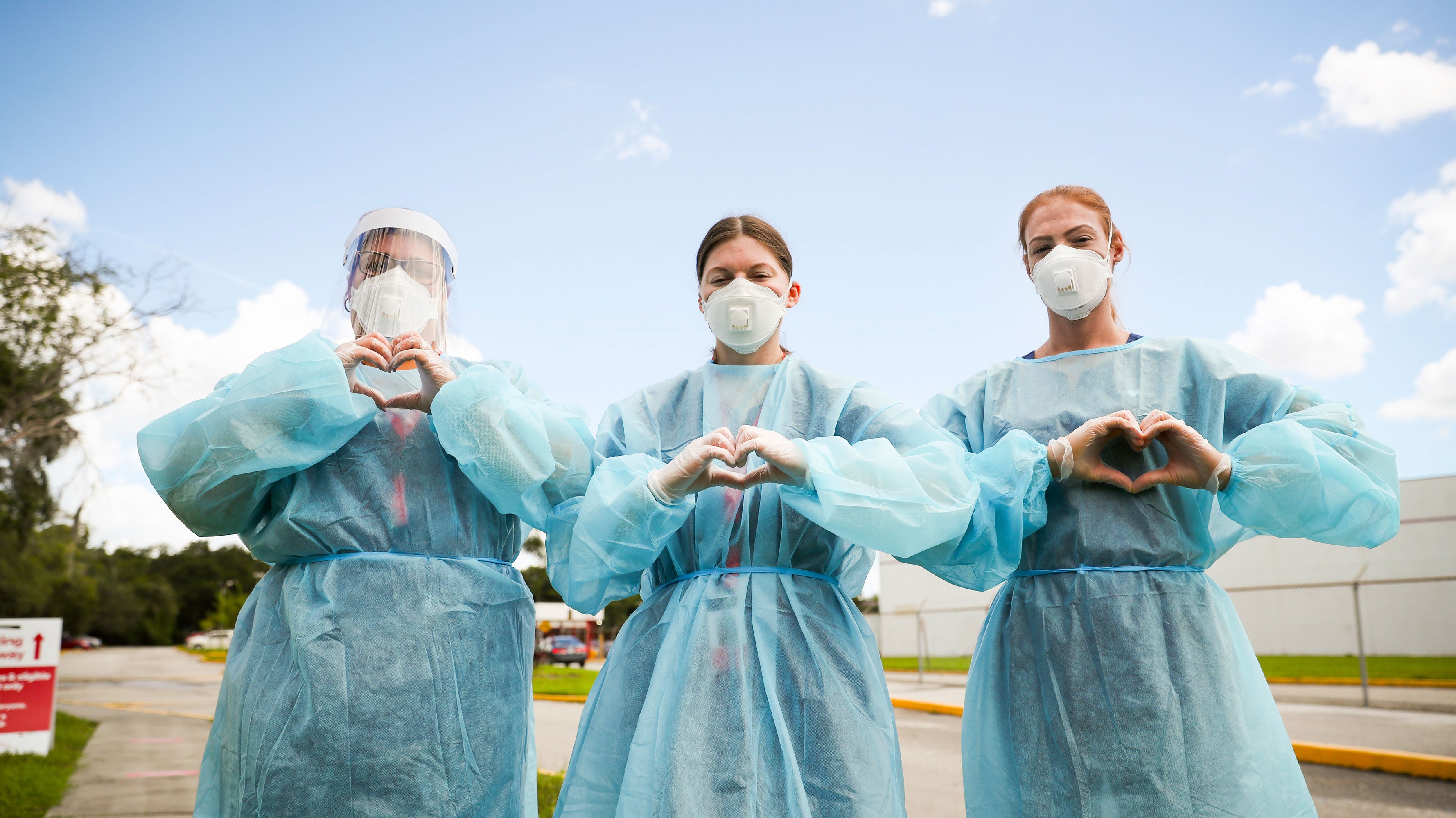 Three health care workers, wearing PPE, make their hands into the shape of hearts.
