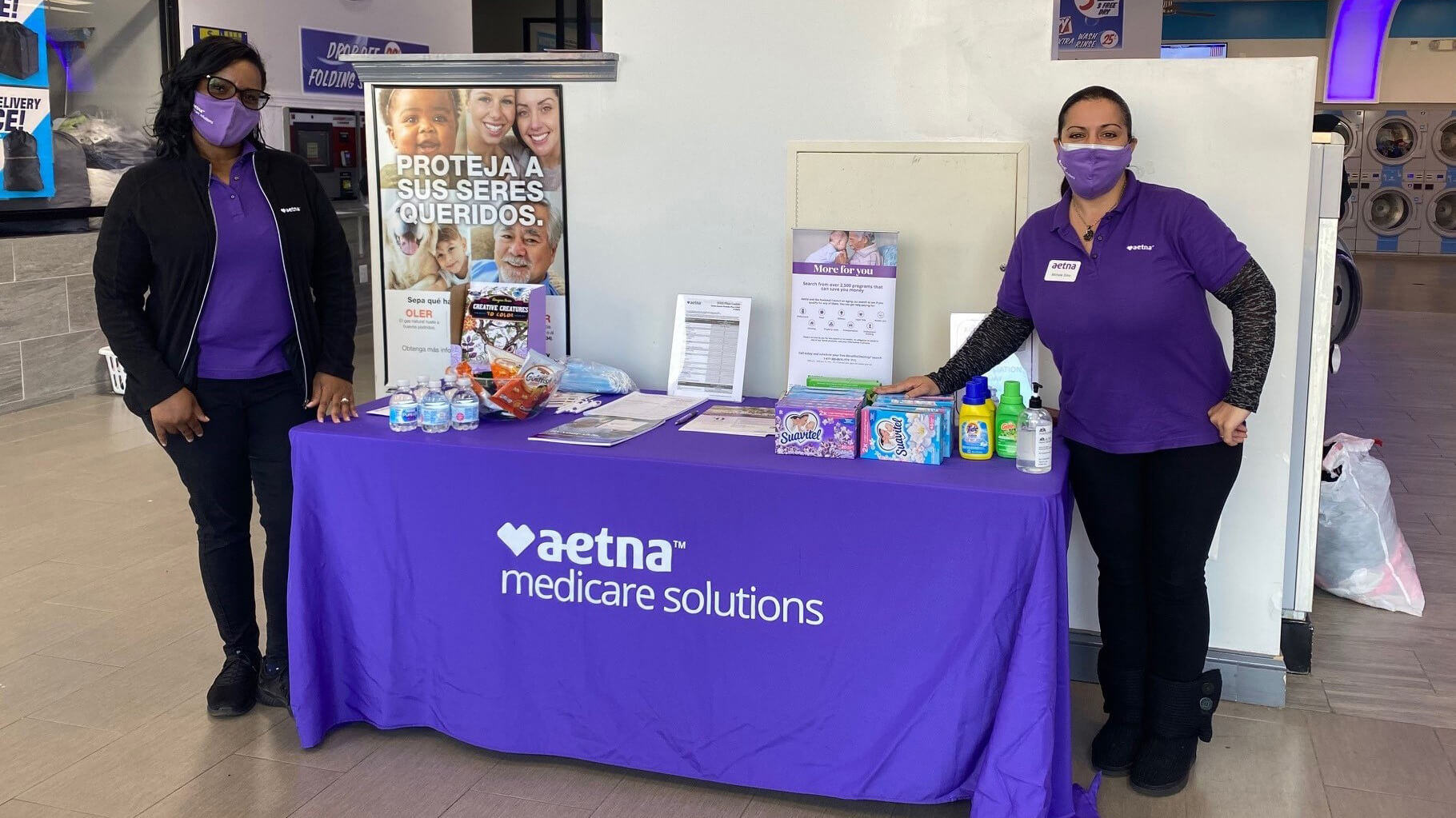 Two masked Aetna representatives provide free laundry products from a table stocked with products.
