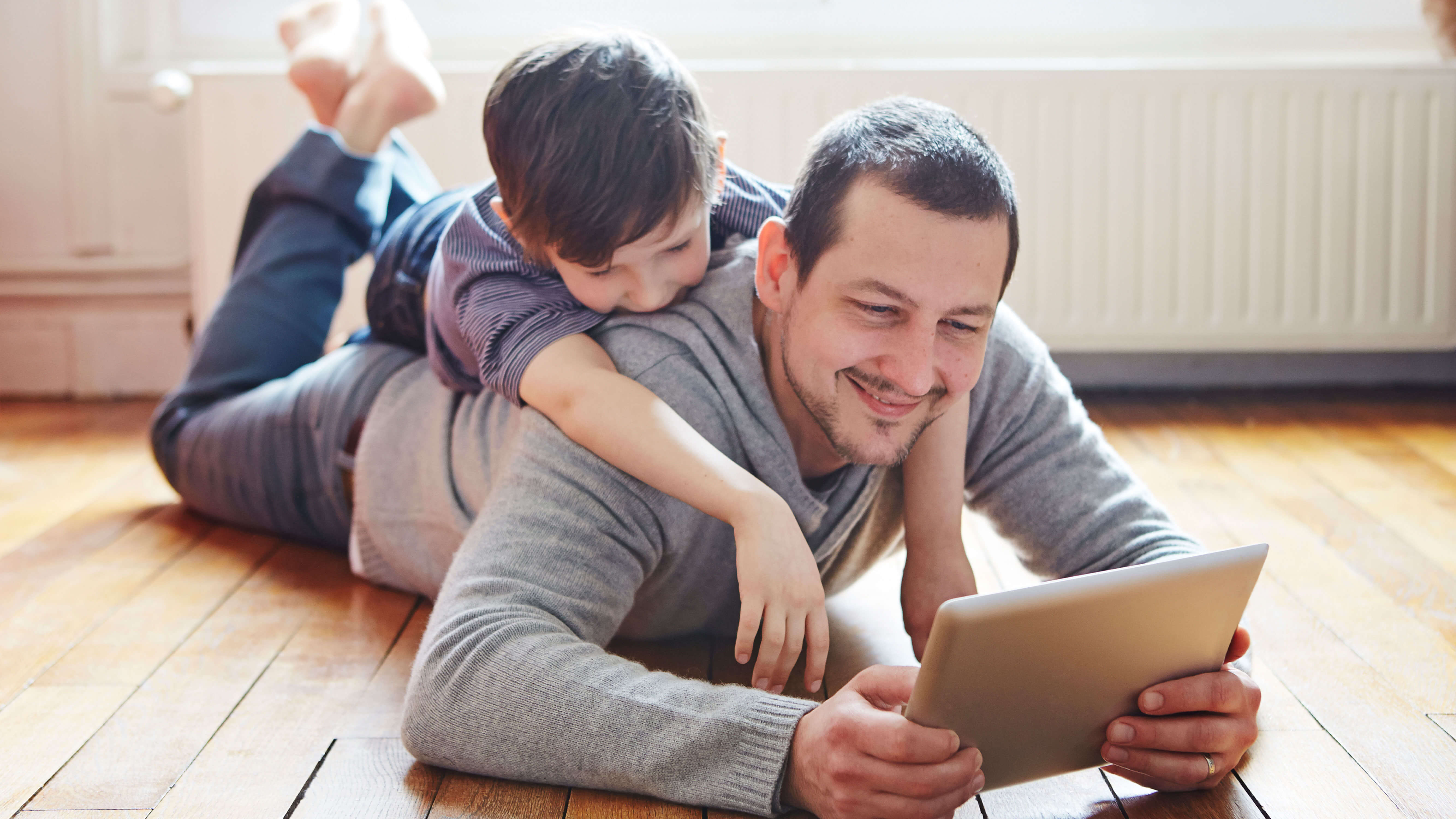 A young father lays on the floor to use his tablet while his son climbs on his back.