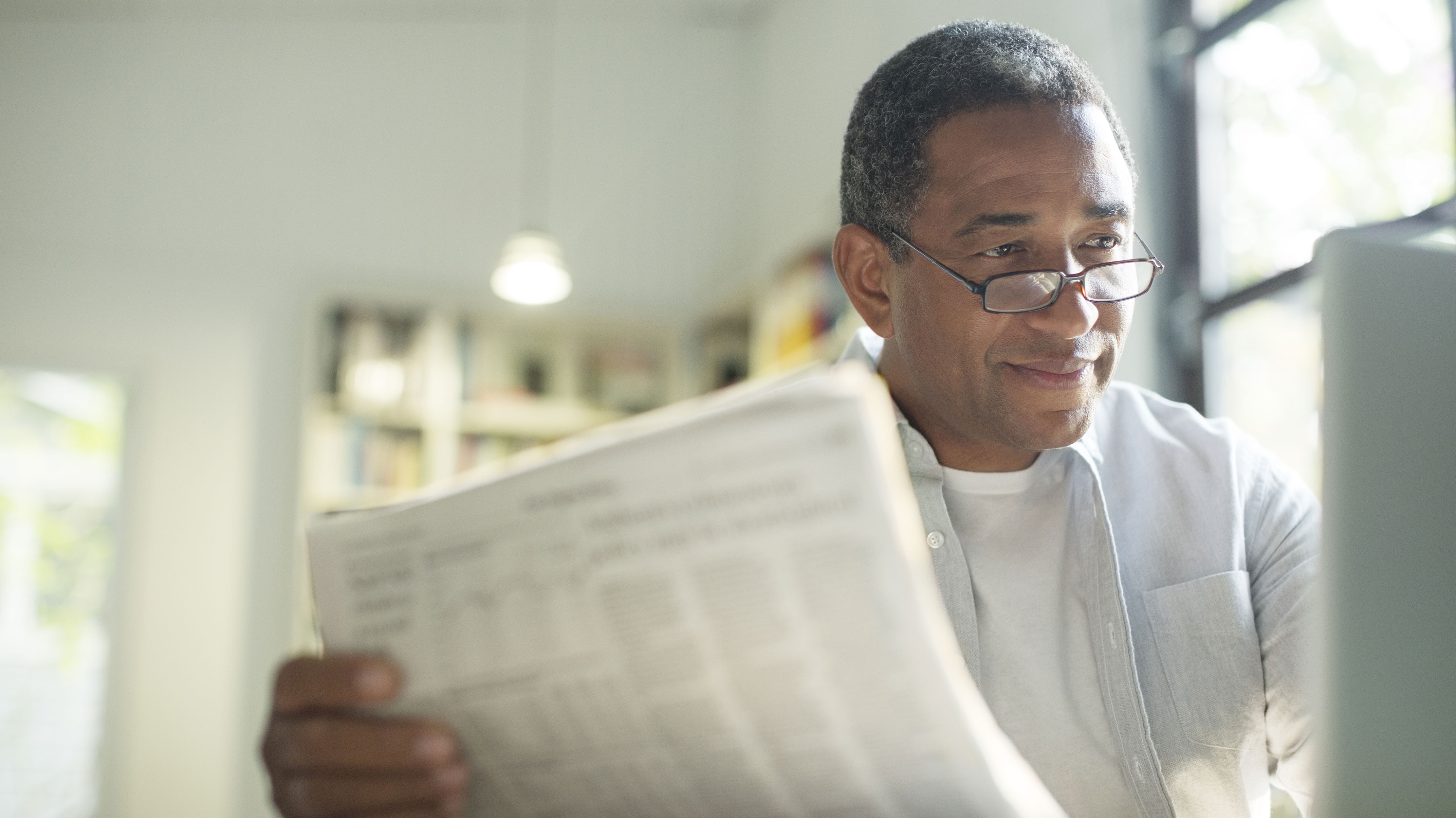 A middle aged man sitting alone at home looking at a computer and holding papers. 