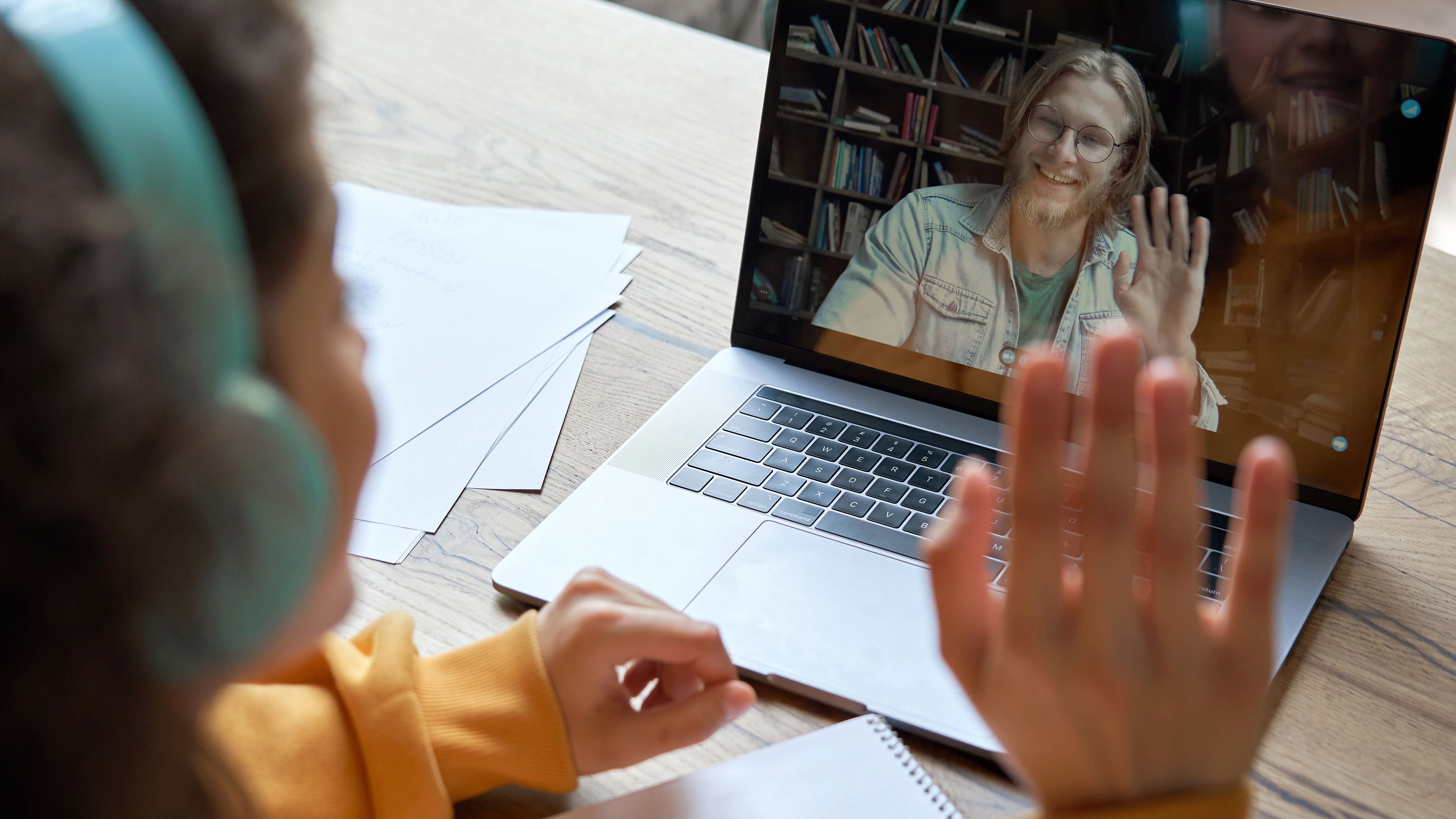 A young man having a video call on a laptop waving at the person he is conversing with. 