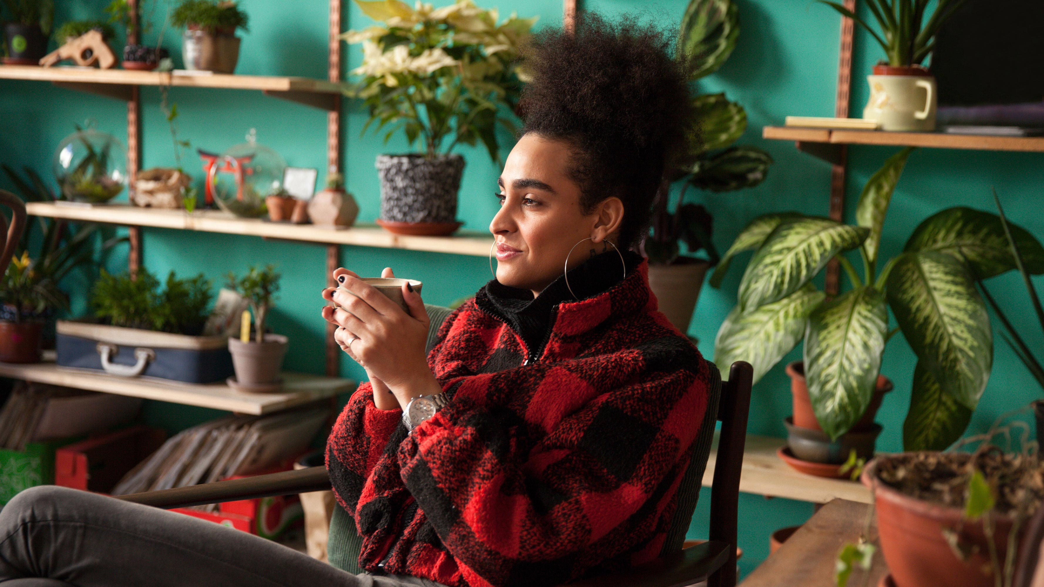 A woman, wearing a red and black sweater, holds a cup of tea with both hands while relaxing in her plant-filled living room.
