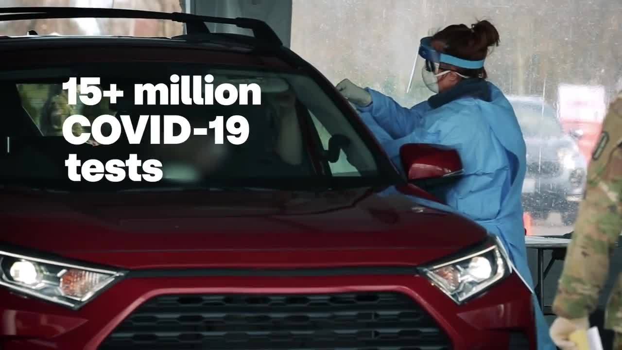 A health care worker, wearing full PPE, reaches through the window of a red car to administer a COVID-19 test.