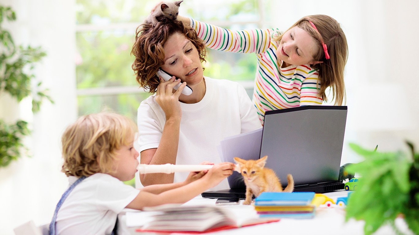 A mother uses her phone and laptop while her young children play on the kitchen table with books, instruments, and kittens.