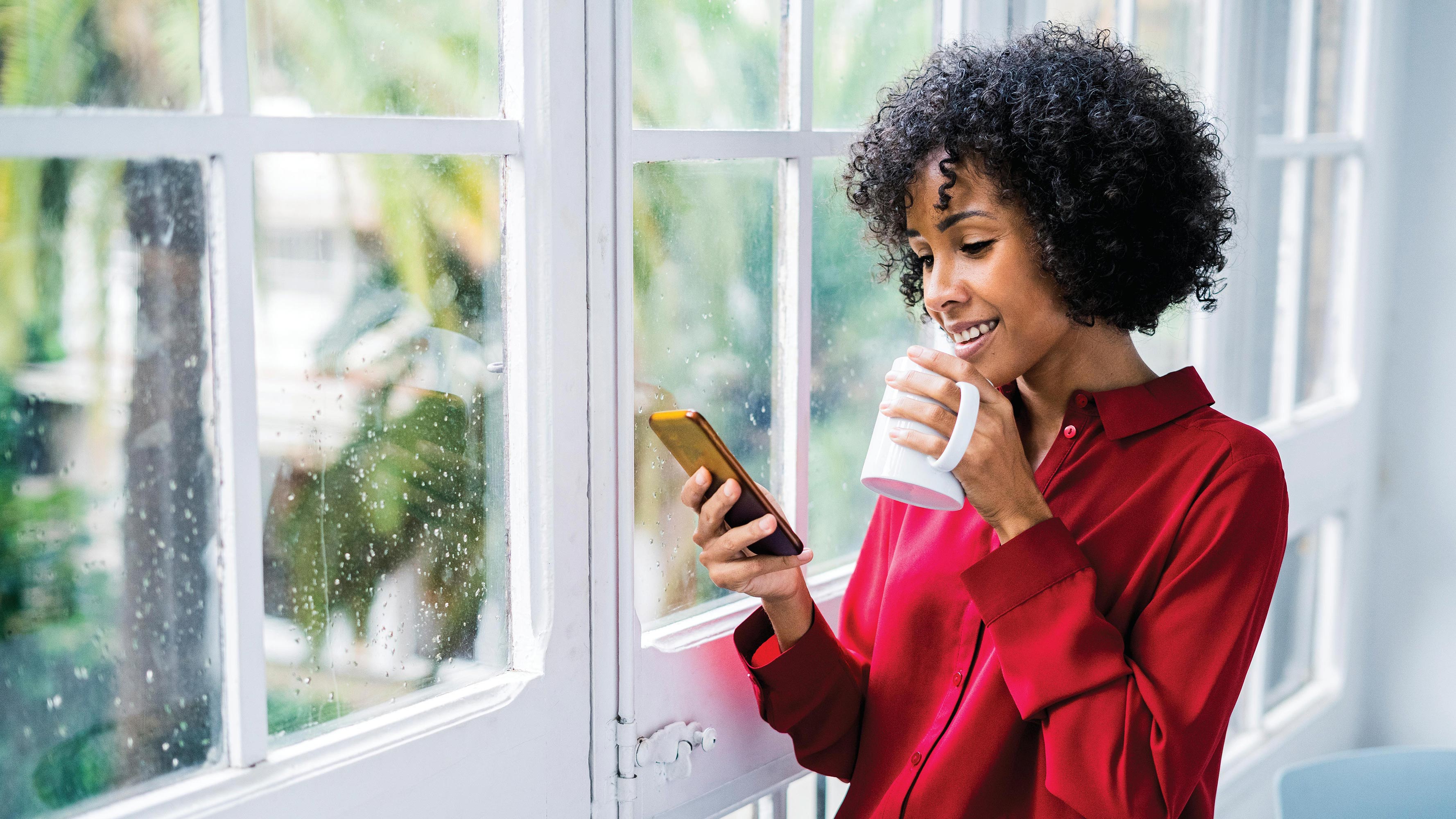 A young woman with curly hair, wearing a red button-down, collared shirt, stands by her window to drink coffee and scroll through her phone.