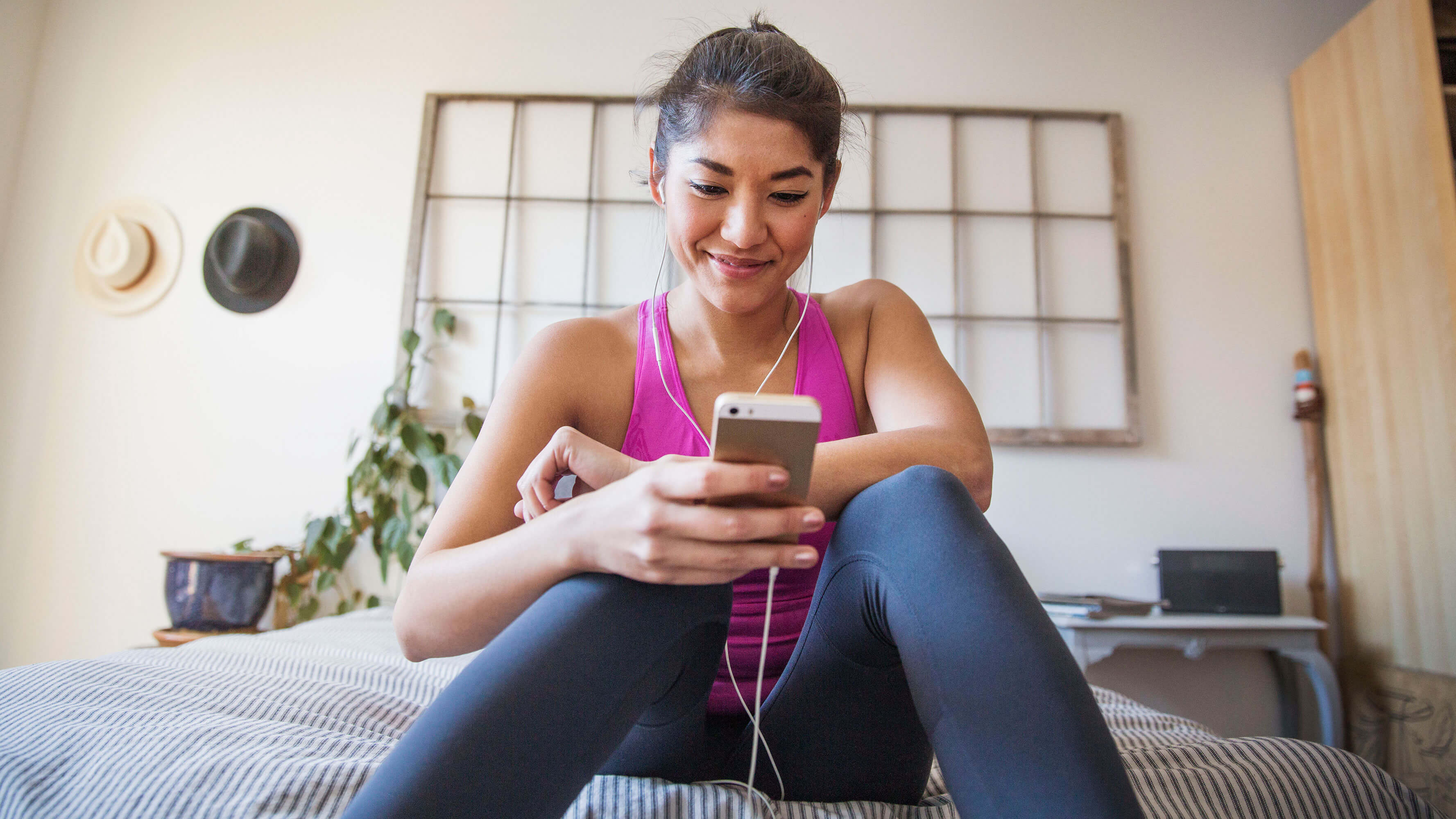 A young woman seated on the edge of her bed listening to a podcast on her phone.