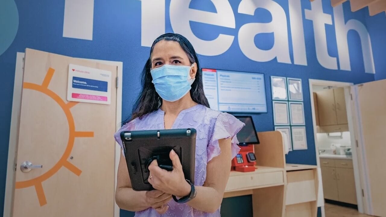 A healthcare provider in blue scrubs with a face mask holds a tablet in the lobby of a HealthHub location.
