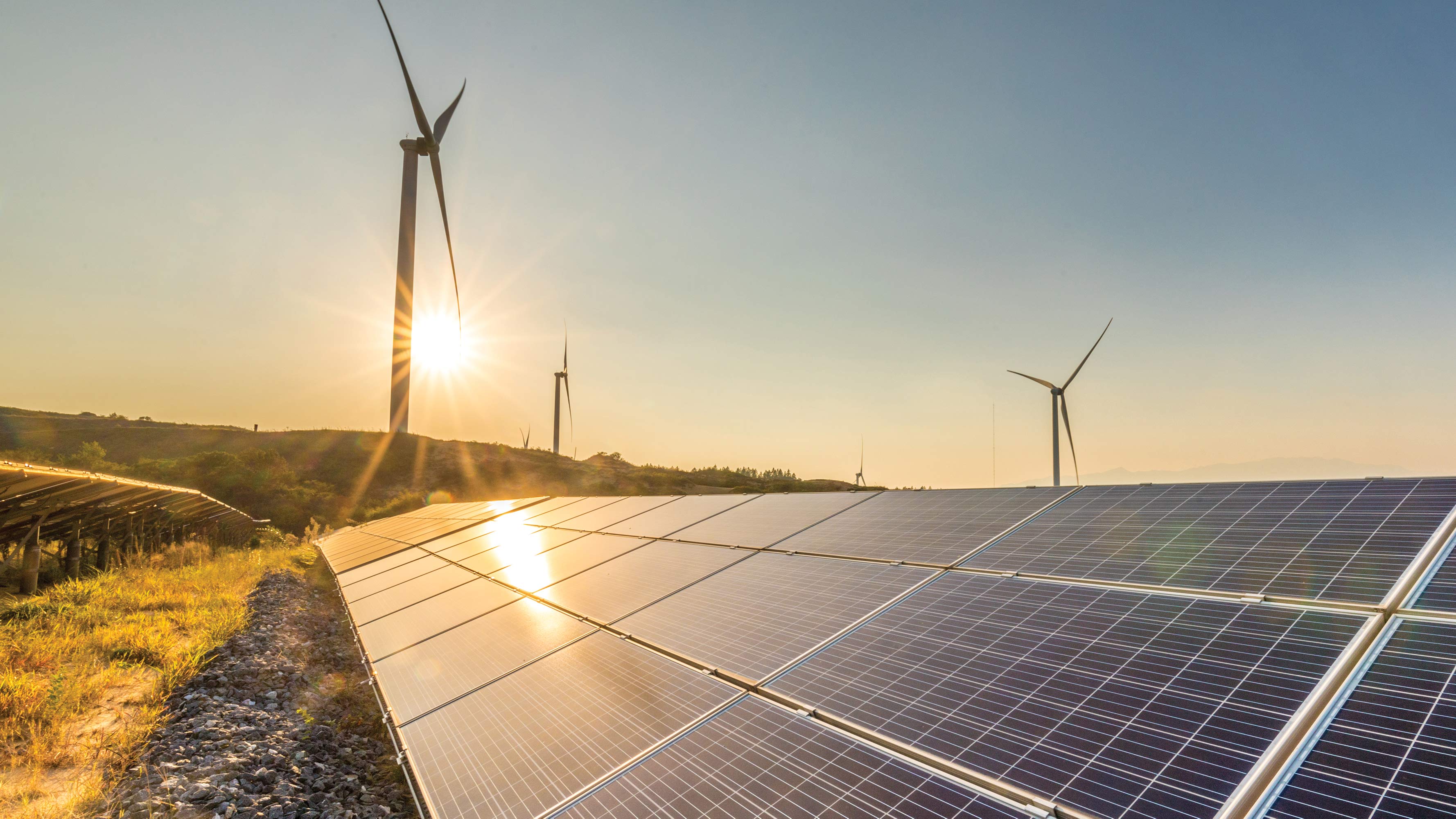 The sun sets behind a green energy field featuring solar panels and wind turbines.