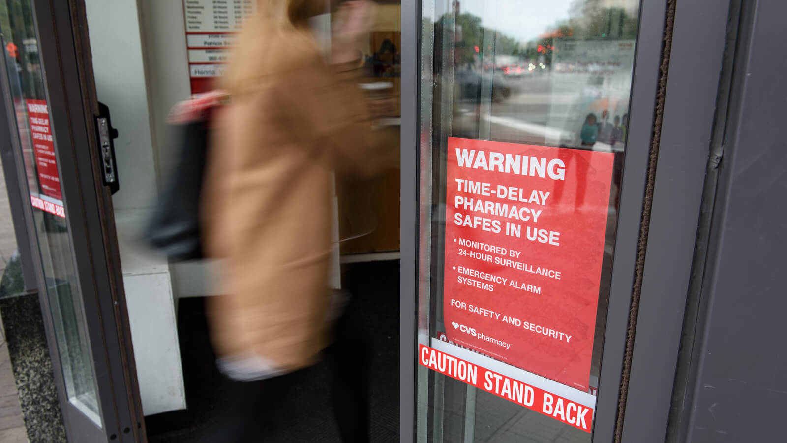 A woman enters a CVS Pharmacy location. A Sign on the door reads &quot;Warning: Time-Delay Pharmacy Safes In Use. Monitored by 24-Hour Surveillance. Emergency Alarm Systems. For Safety And Security. CVS Pharmacy&quot;