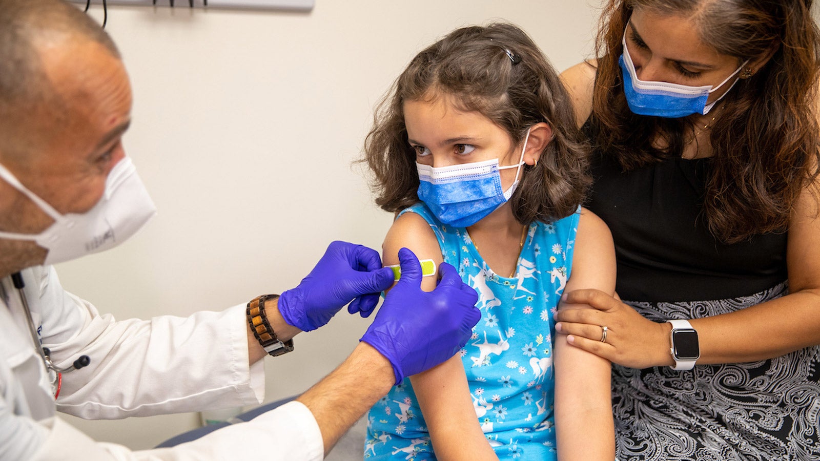 A healthcare provider puts a bandage on a young patient&#039;s arm while her mother comforts her.