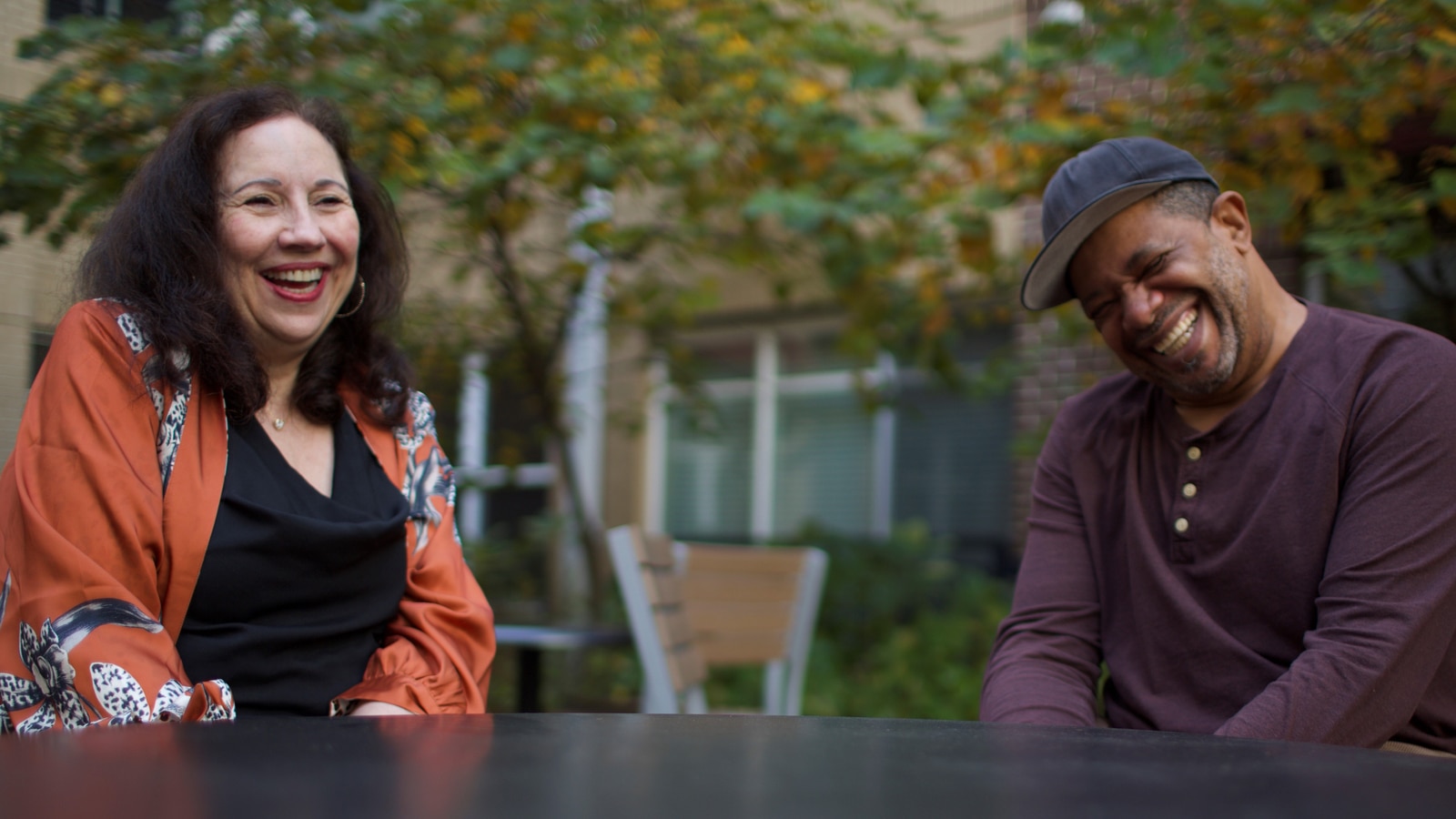 Frances Lopes and Javier Edwards laugh together as they sit at a table outdoors.