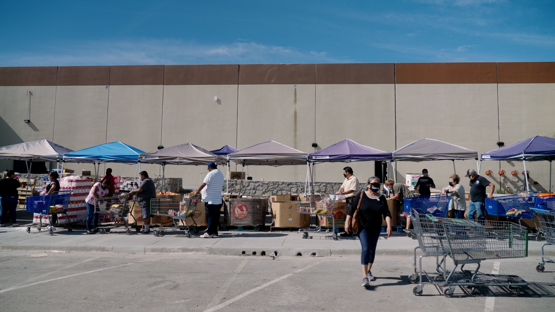 A group of people with shopping carts are lined up in front of a series of tents containing food items.