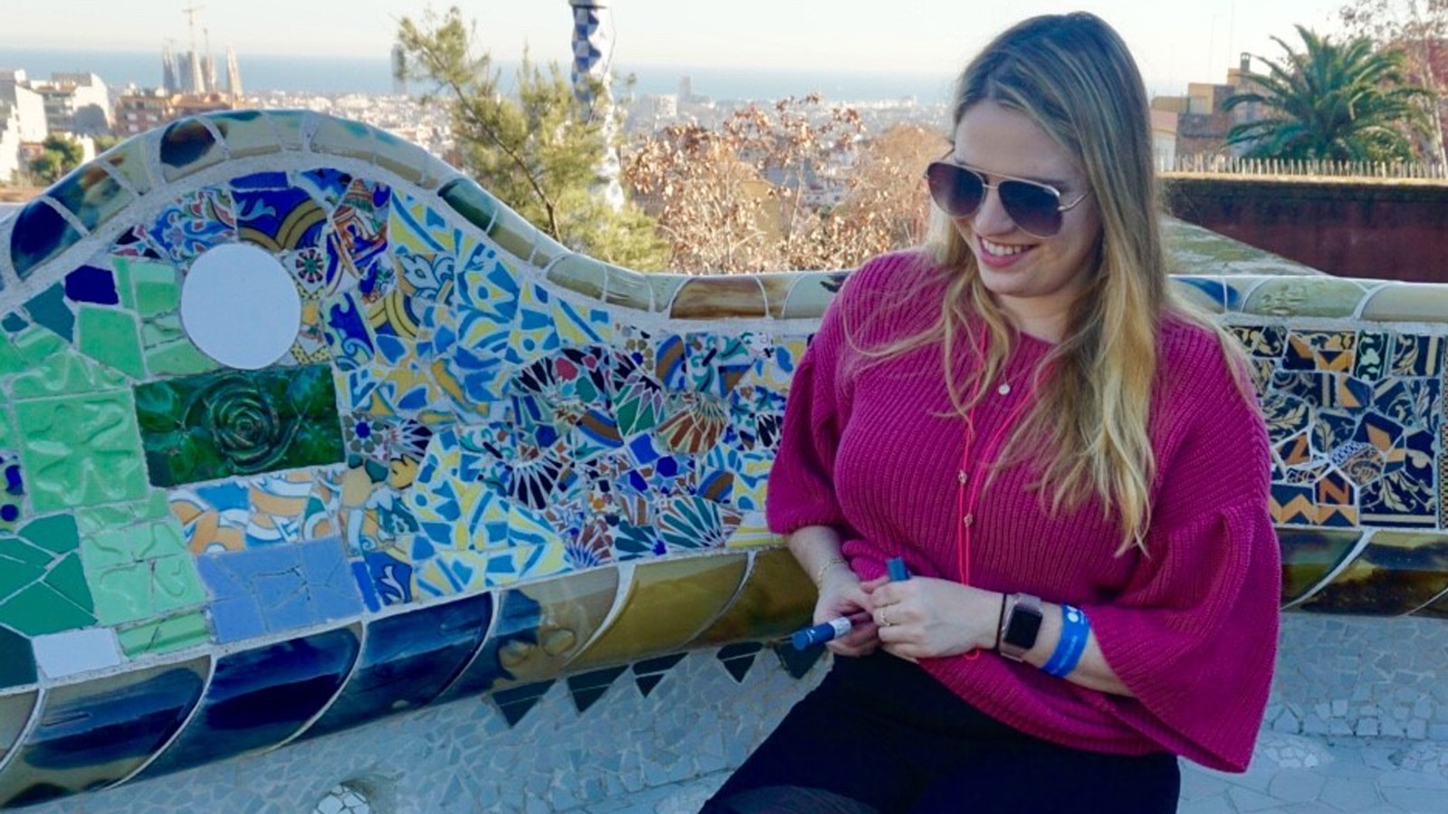 A woman with sunglasses, who is sitting outside, holds a self-injecting medication pen in her hands.