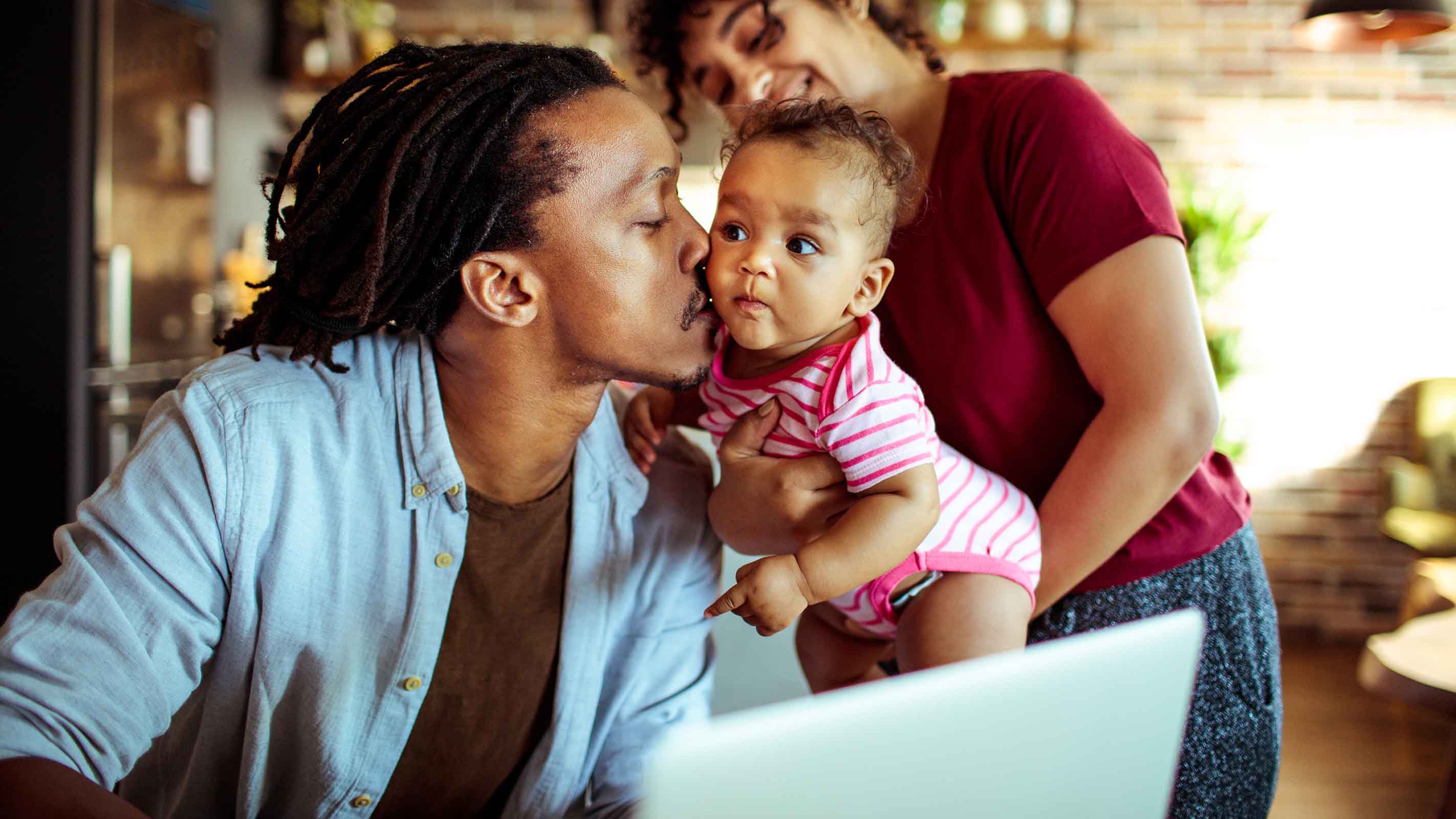 A father leans away from his laptop to kiss his baby daughter while she's held by her mother.