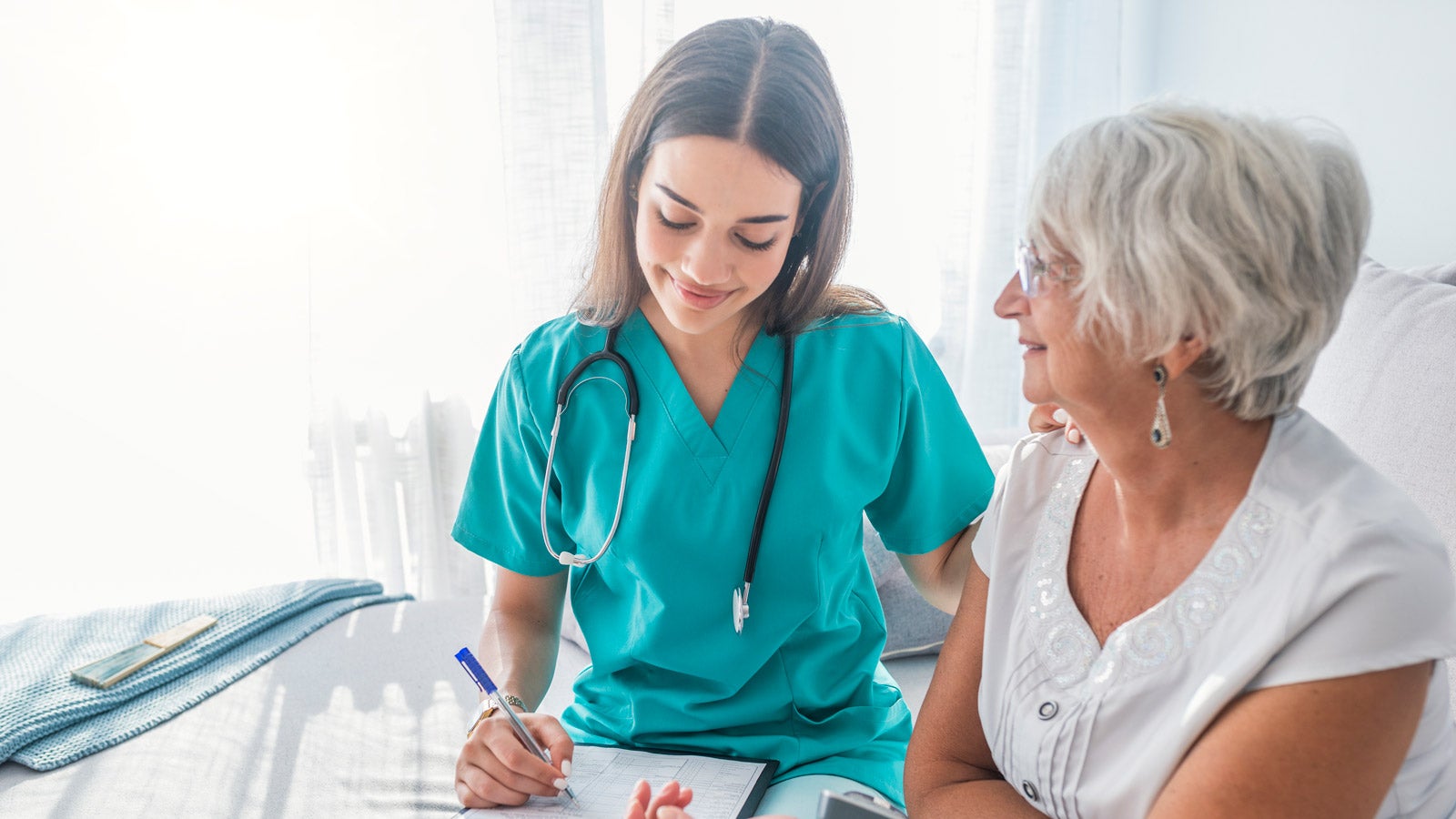 A senior patient smiles, and a healthcare provider fills in a form on a clipboard.
