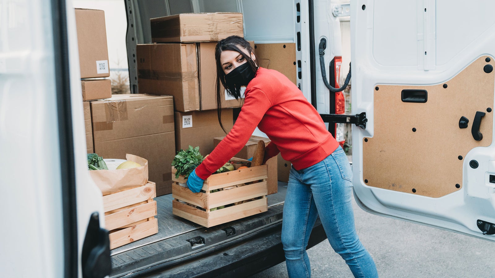 A masked and gloved volunteer carries a wooden box filled with produce.