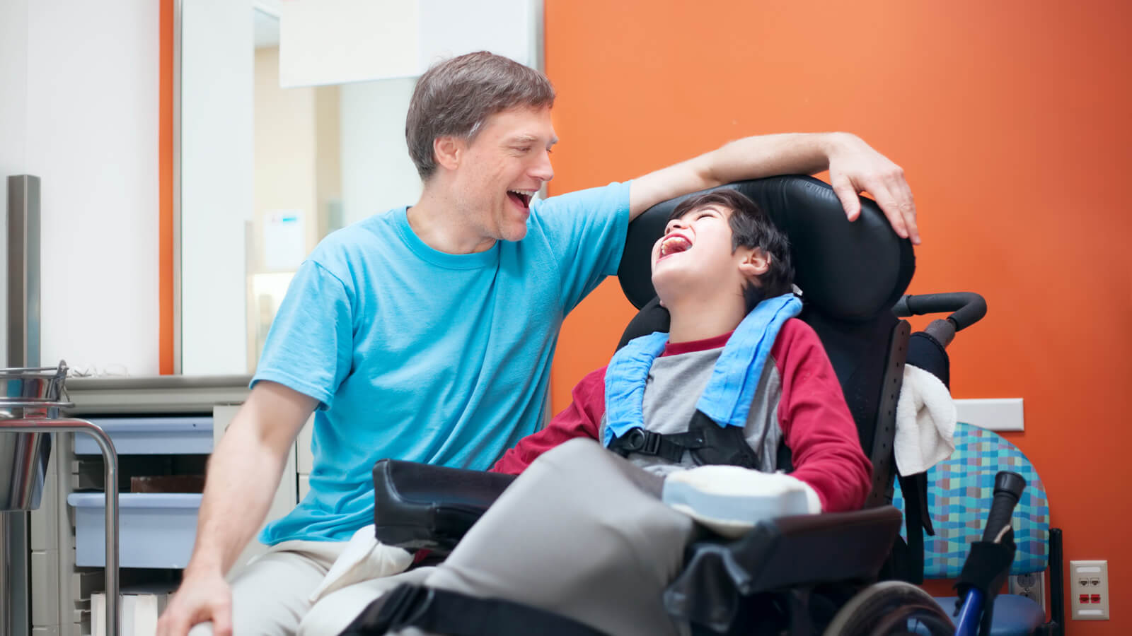 A caregiver and a young boy in an electric wheelchair smile at each other.