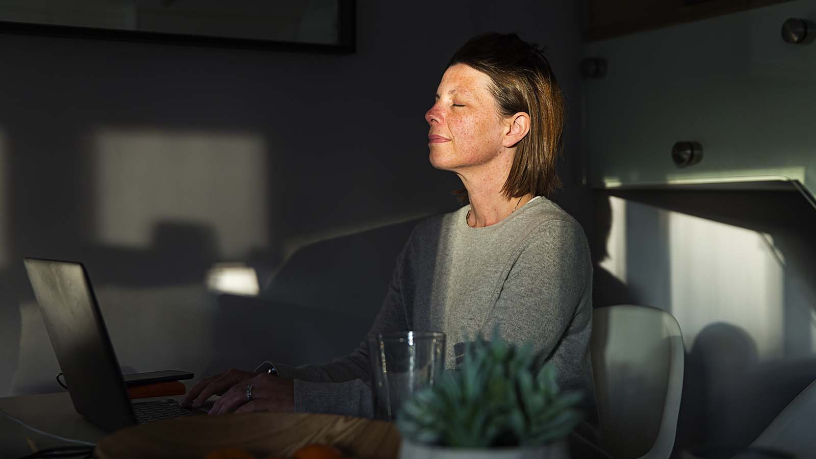 A woman sits at a dining table with laptop, taking a pause to enjoy the sunshine.