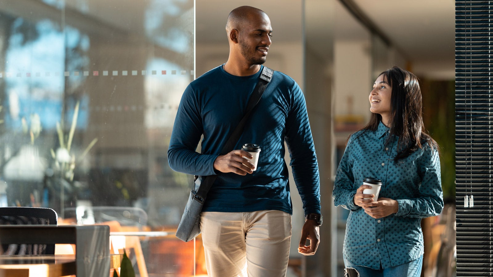 Two smiling colleagues with disposable coffee cups walk together.