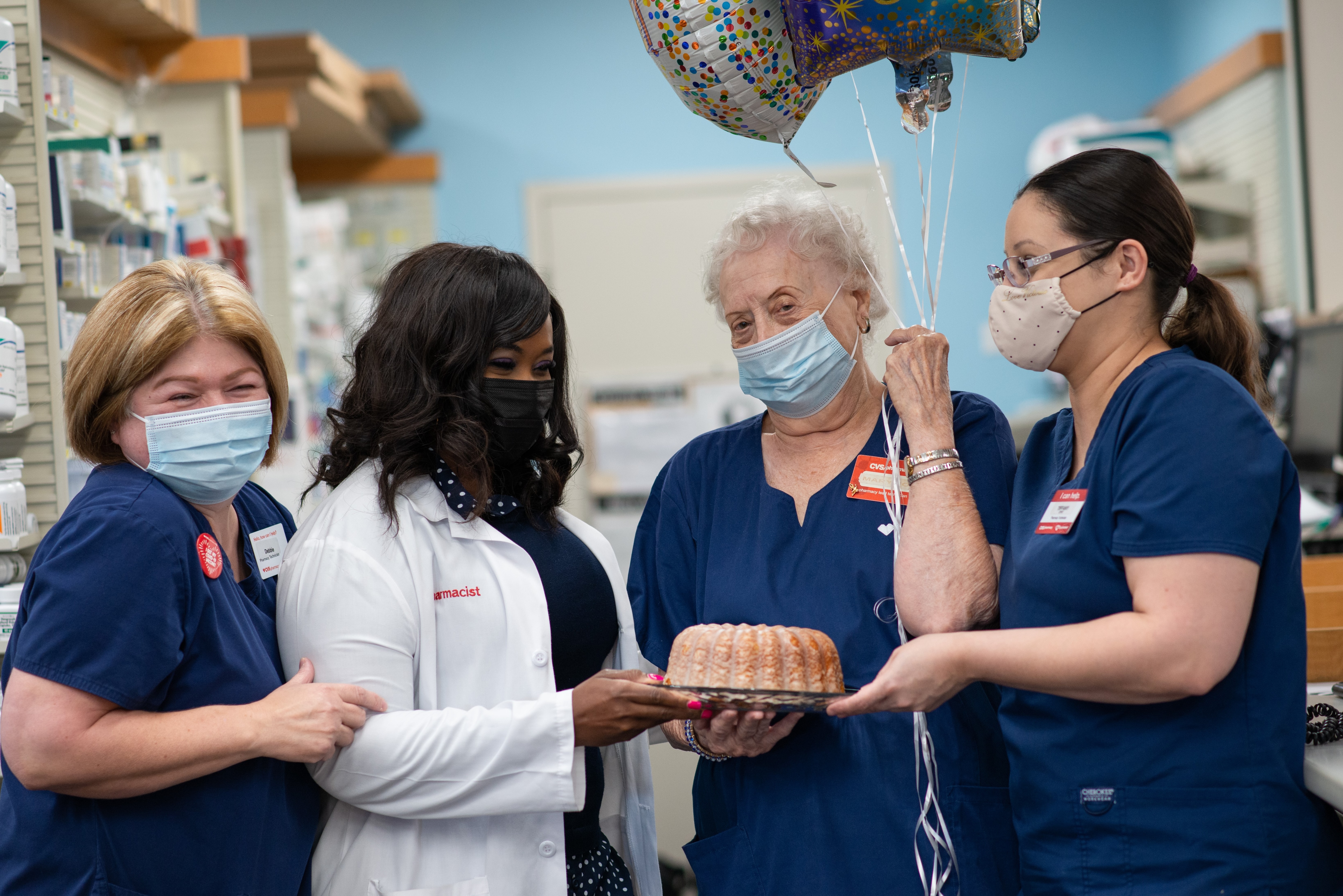 Three masked employees celebrate Mary Jo, a long-term CVS Pharmacy employee, with balloons and a bundt cake.