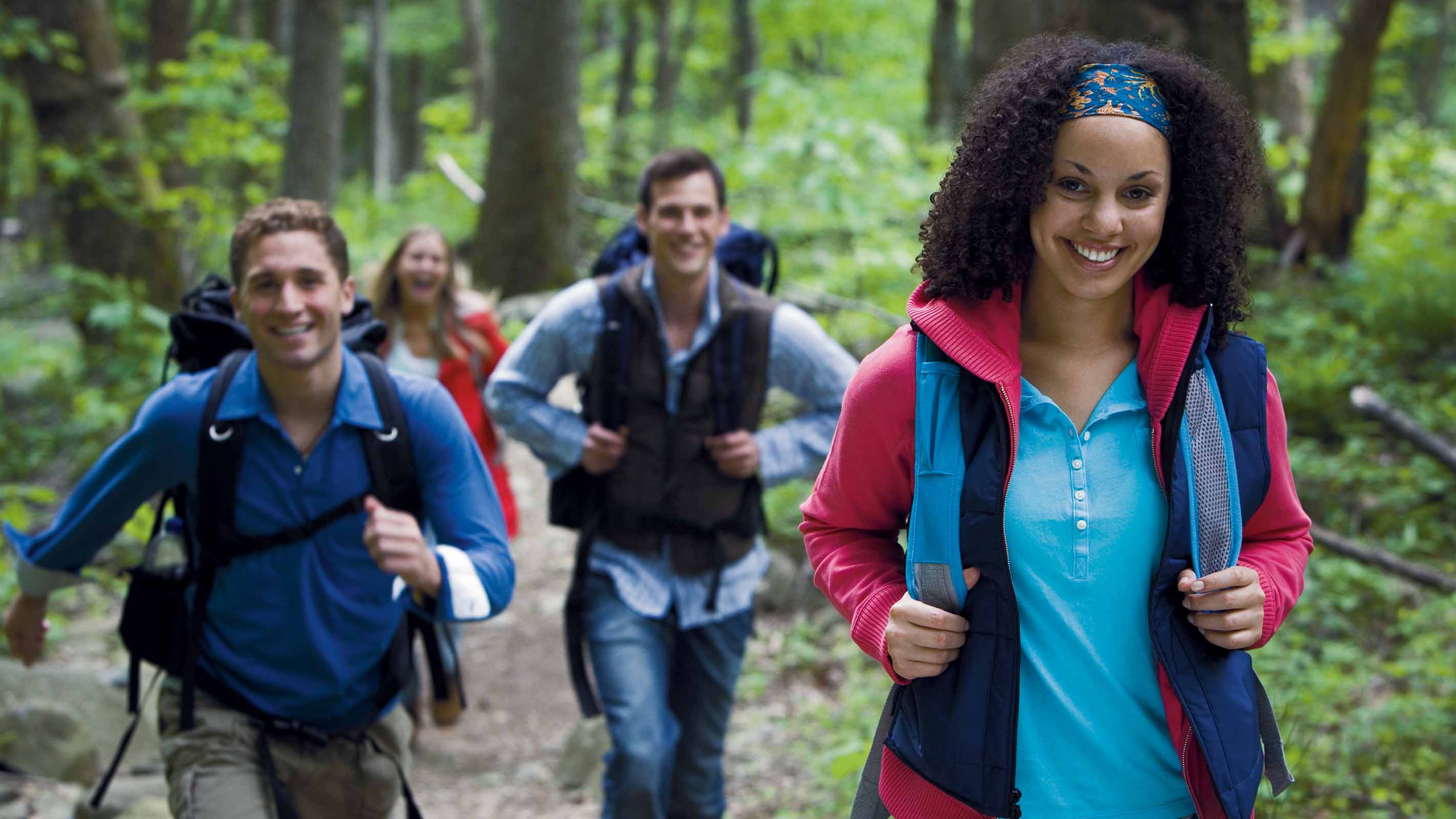 A group of friends smile while hiking through a wooded area, wearing backpacks.