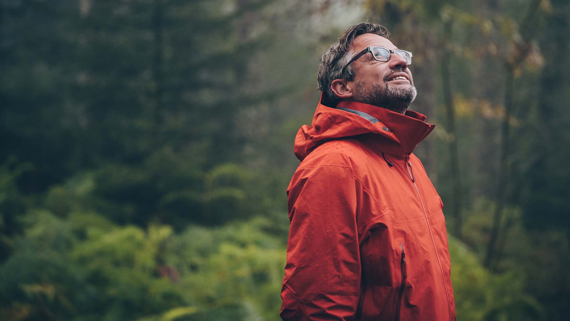 A mean wearing a red all-weather coat looks upward at a forest canopy.