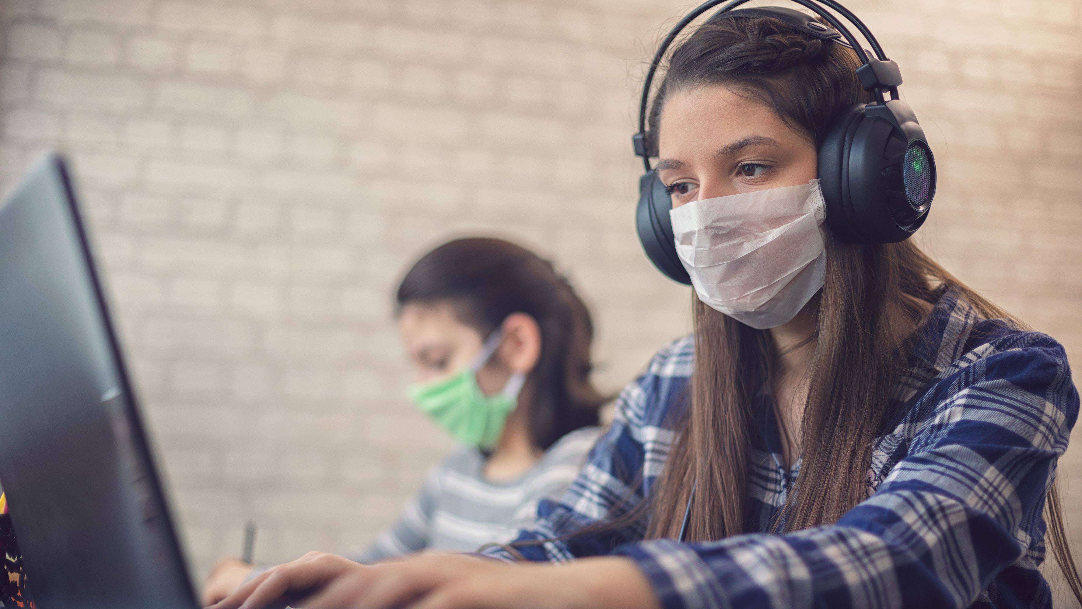 A young female student is seen wearing headphones and a face mask using a computer.