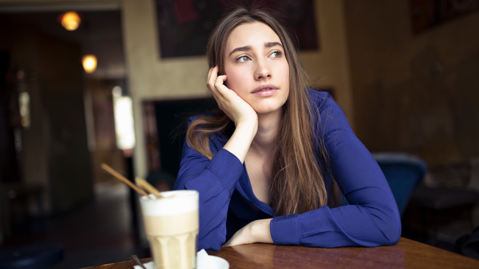 A woman sits at a cafe table looking out and resting her hand on her chin.