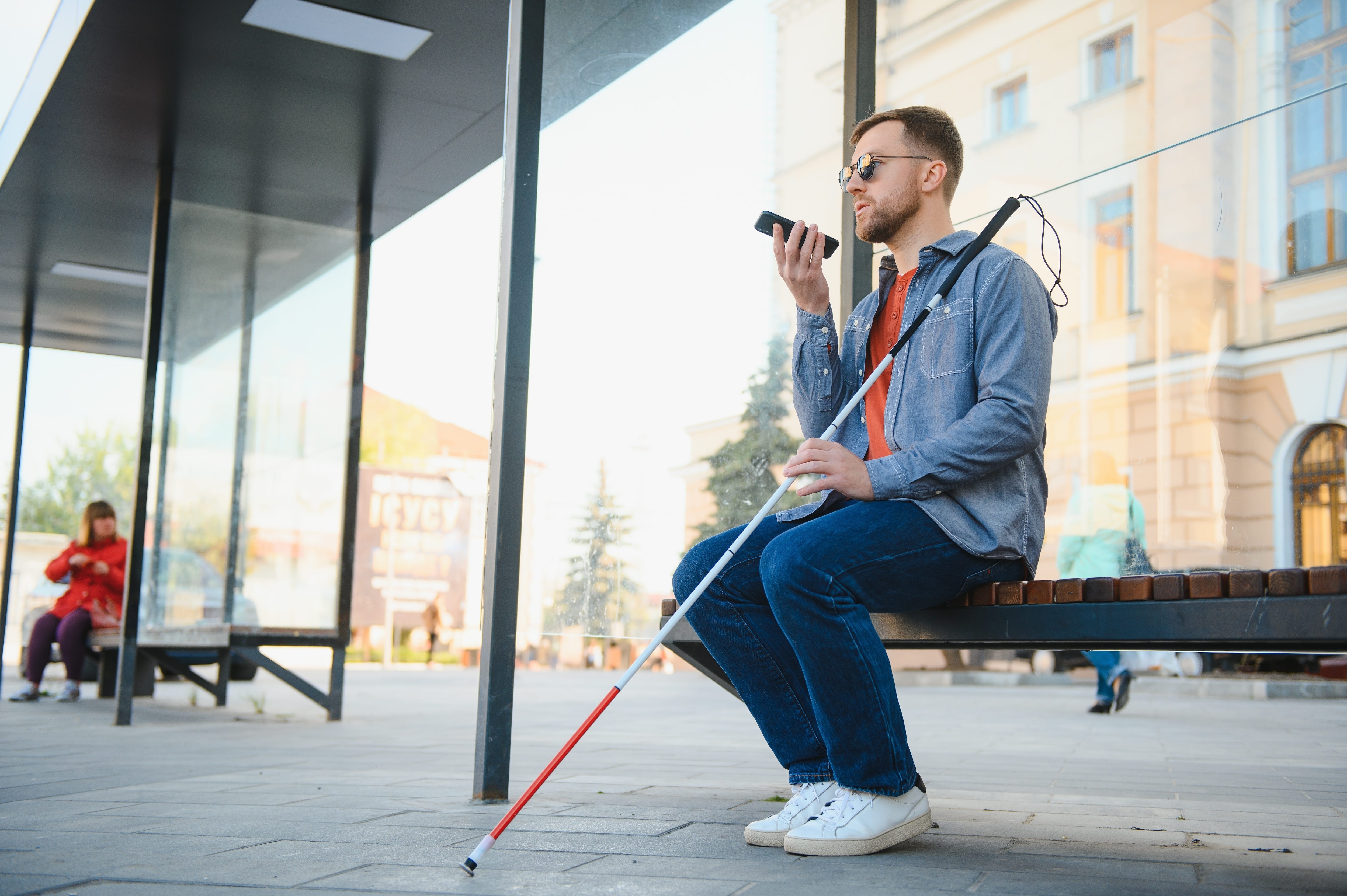 A man with short beard and sunglasses sits on a bench, holding a white cane while speaking into a cell phone.