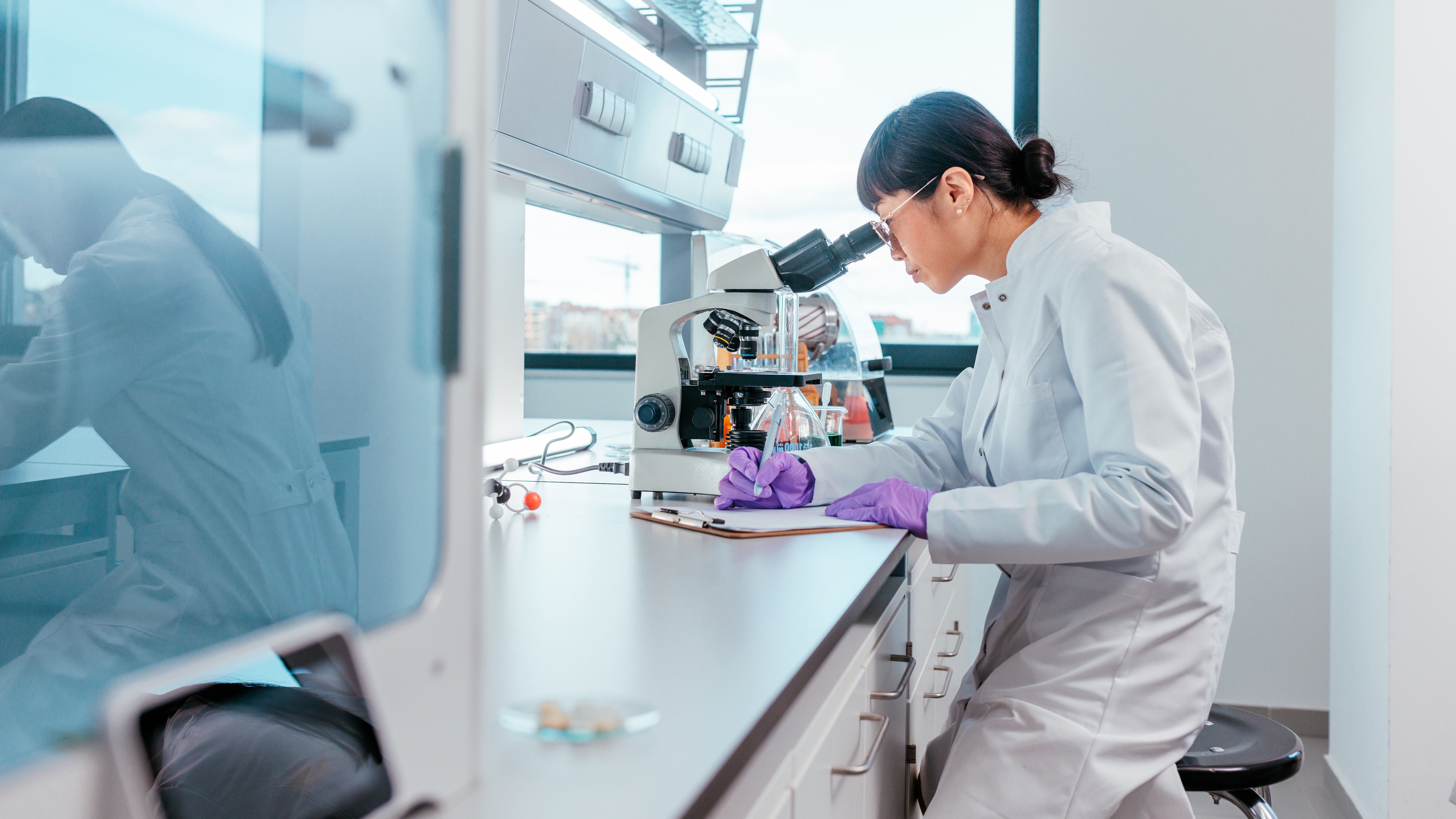 Female scientist in a lab looking in microscope and taking notes
