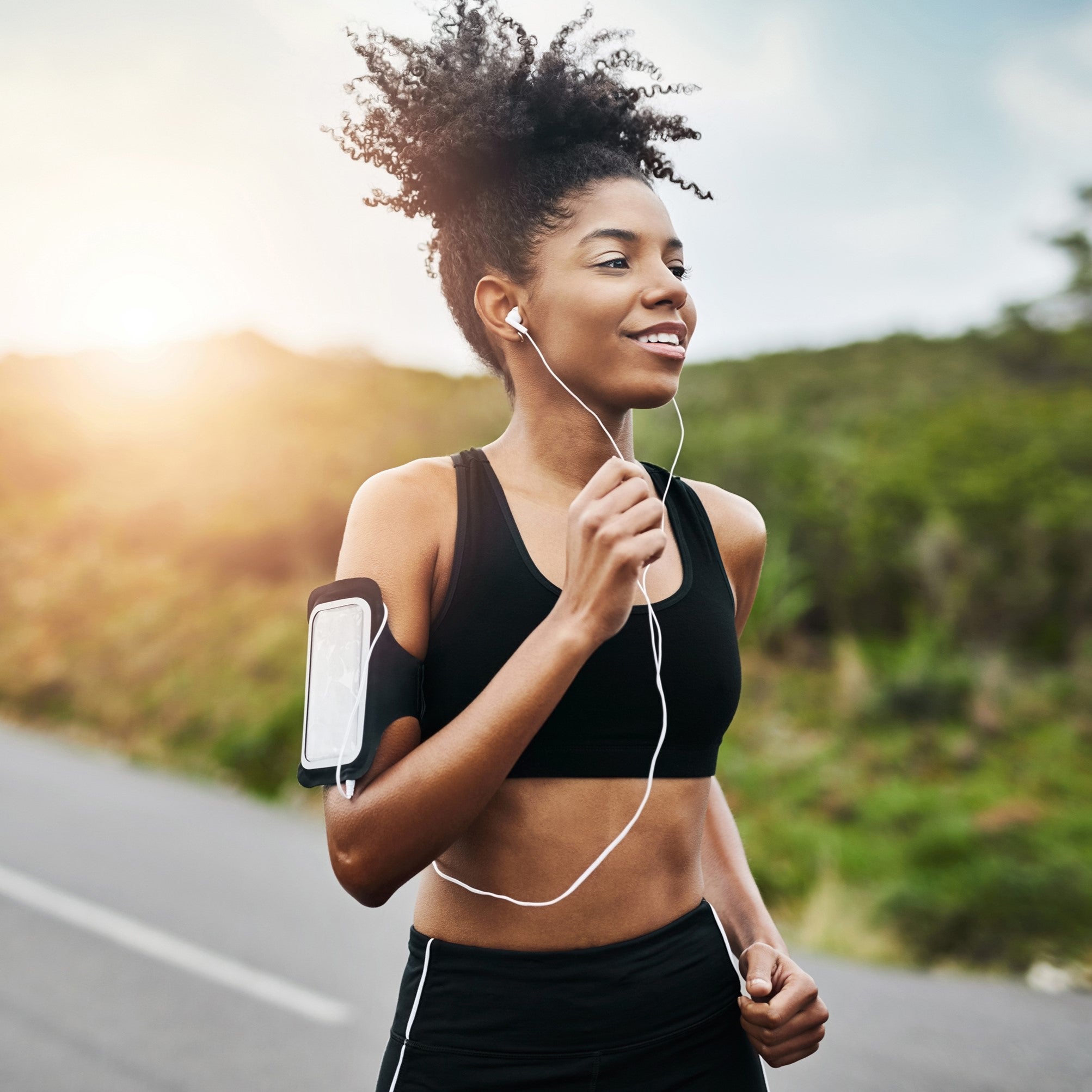 A woman, wearing athletic clothes and headphones, runs on a paved road with her phone strapped to her arm.