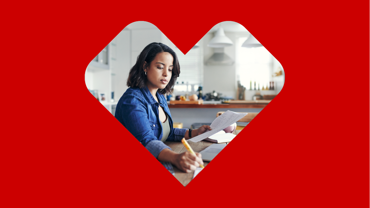 A red overlay of a heart reveals a woman taking notes while working on her laptop at her table.