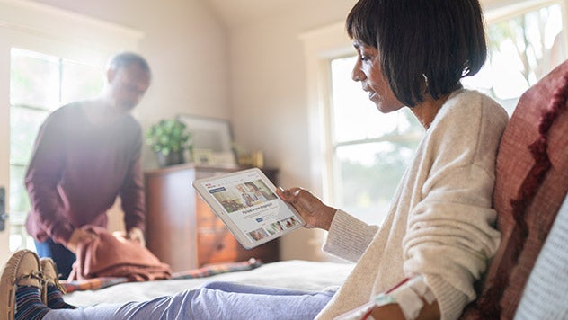 Woman in bed on tablet with male putting clothes away in bedroom