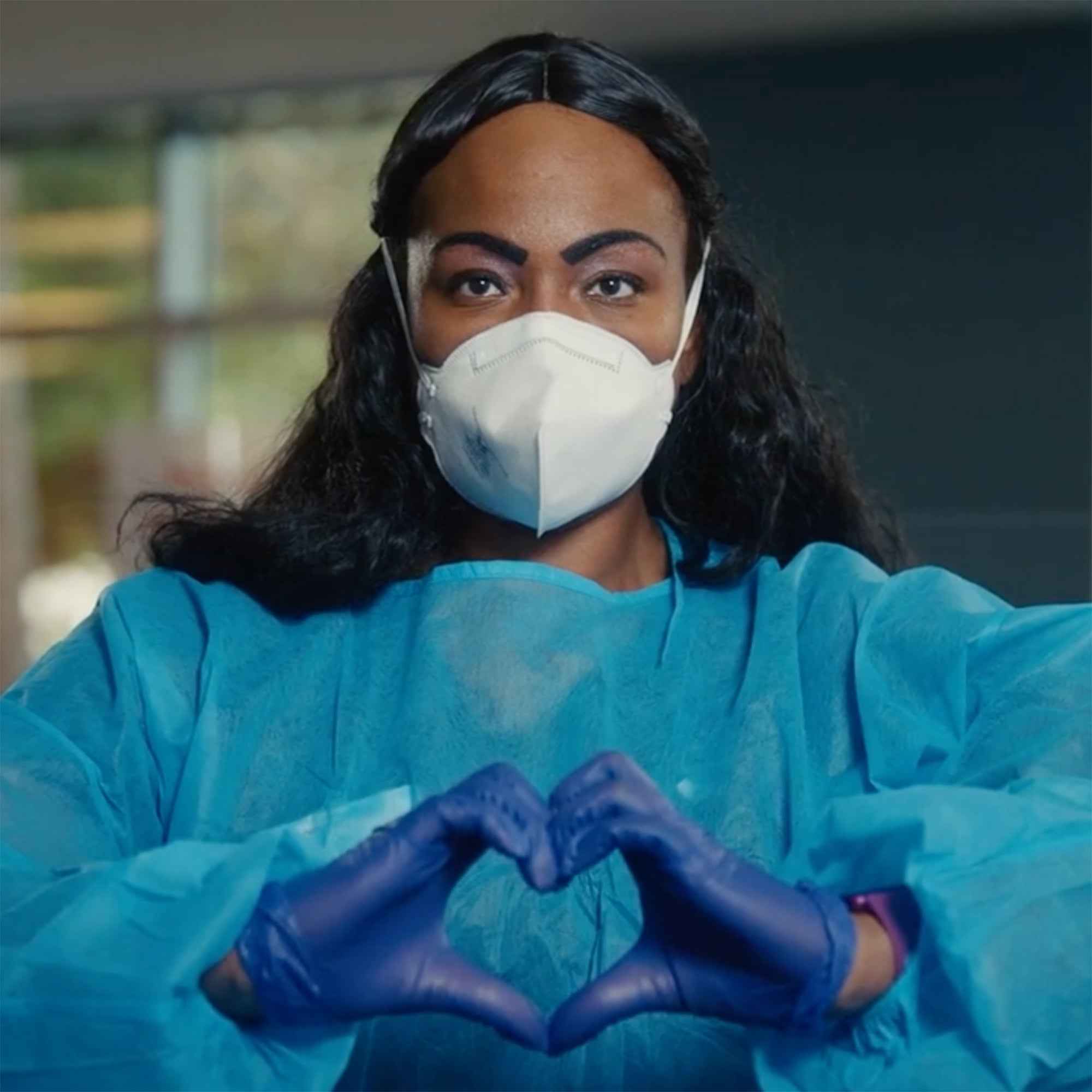 A doctor wearing PPE uses her hands to form the shape of a heart.