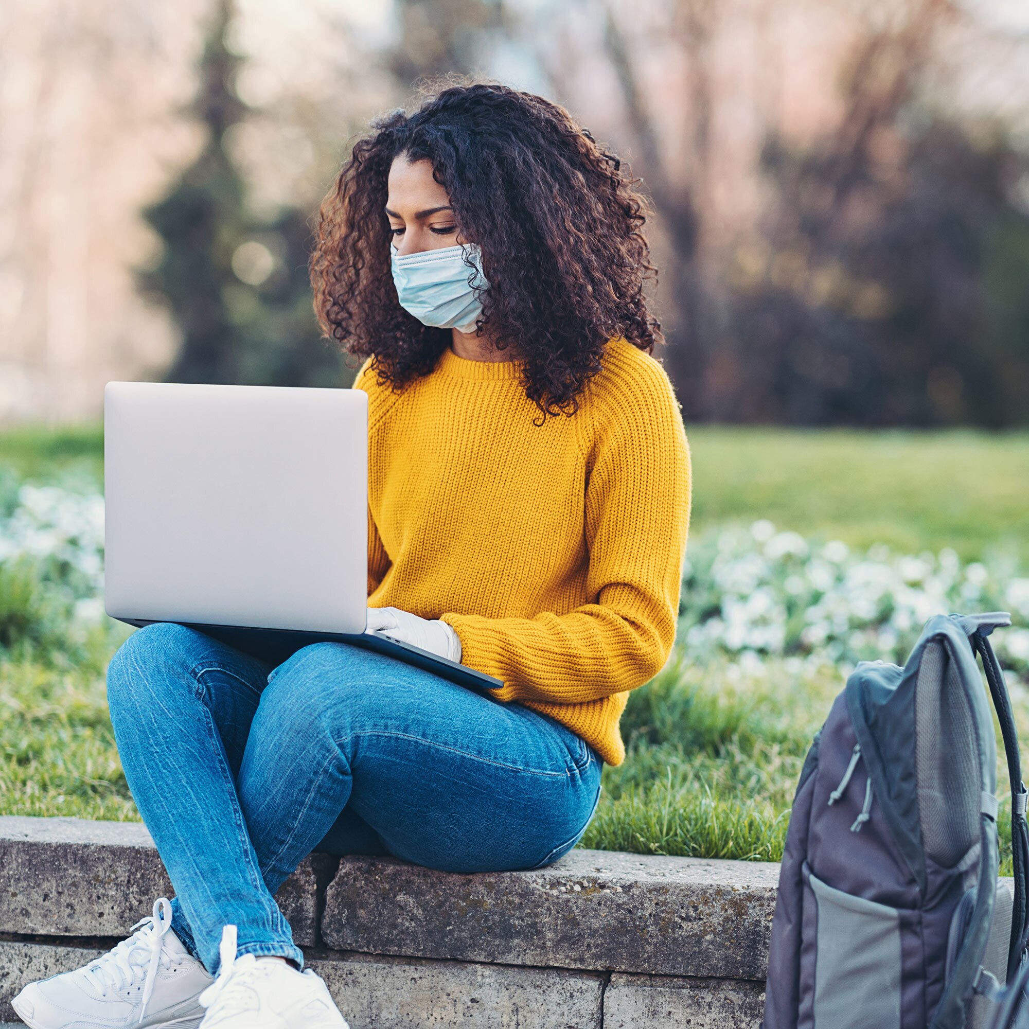 A masked woman wearing a yellow sweater sits in a park to use her laptop.