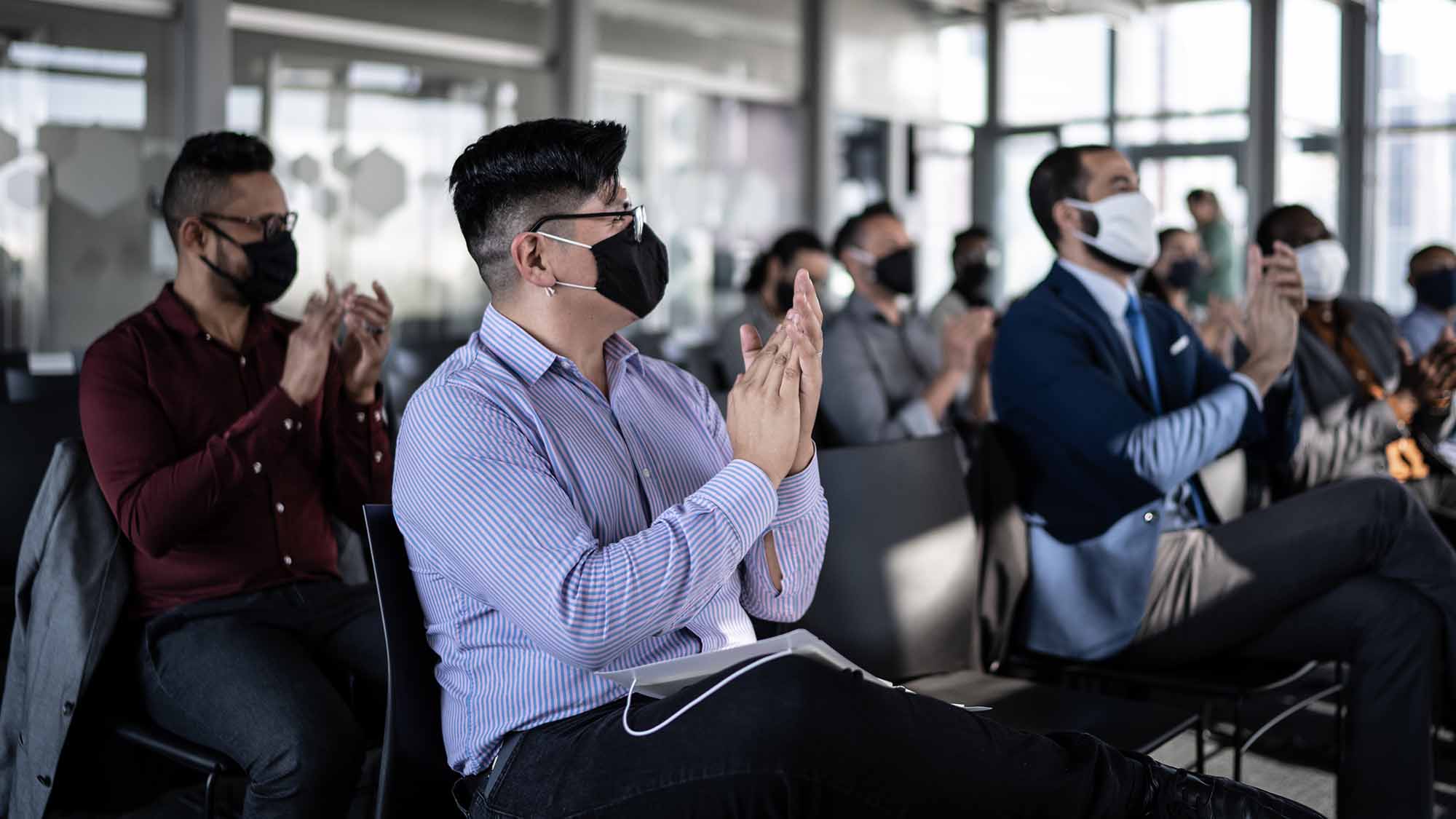 A group of people, wearing face masks, sitting and listening to a presentation in a conference room.