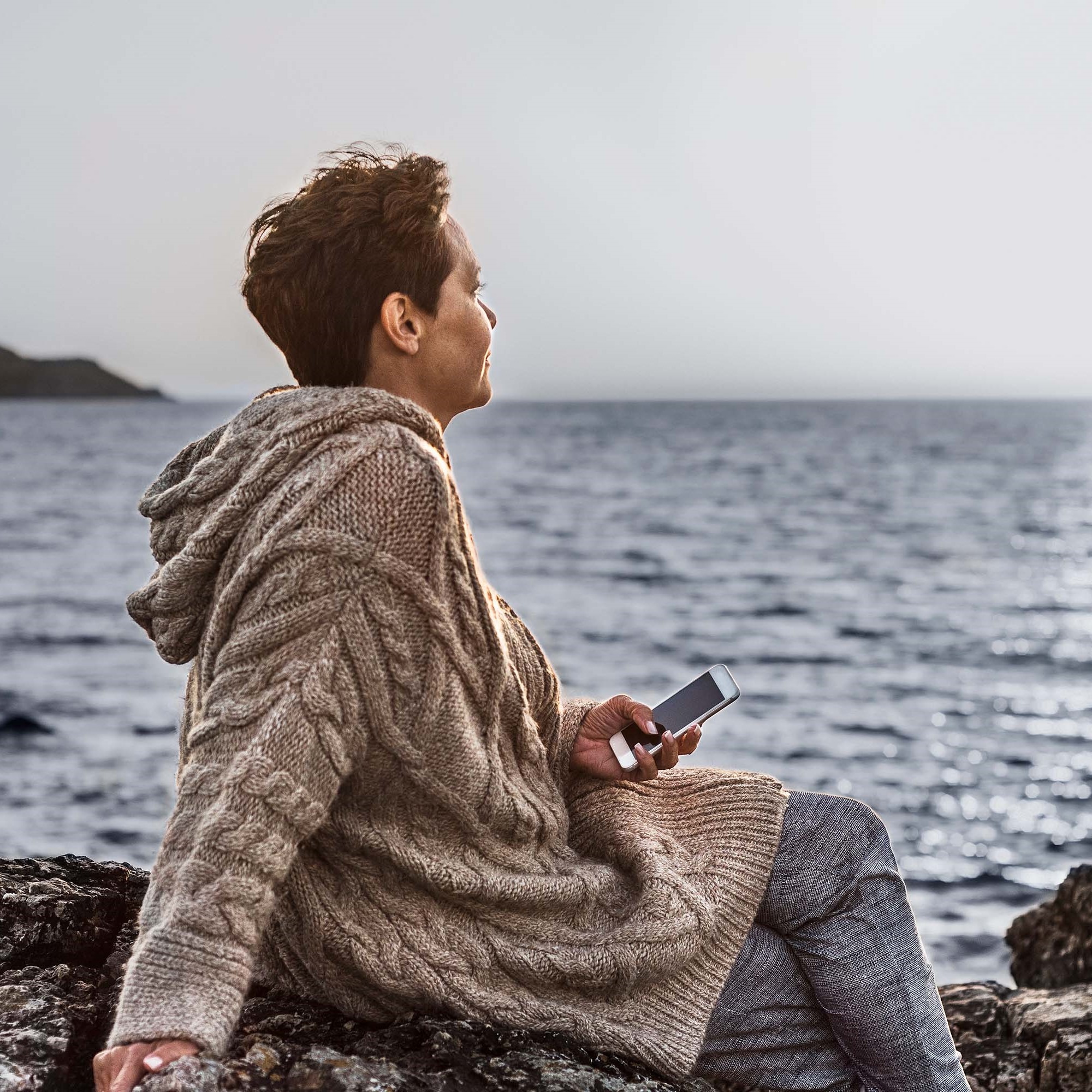 A person sits by the seashore, sitting on rocks, holding a mobile telephone.