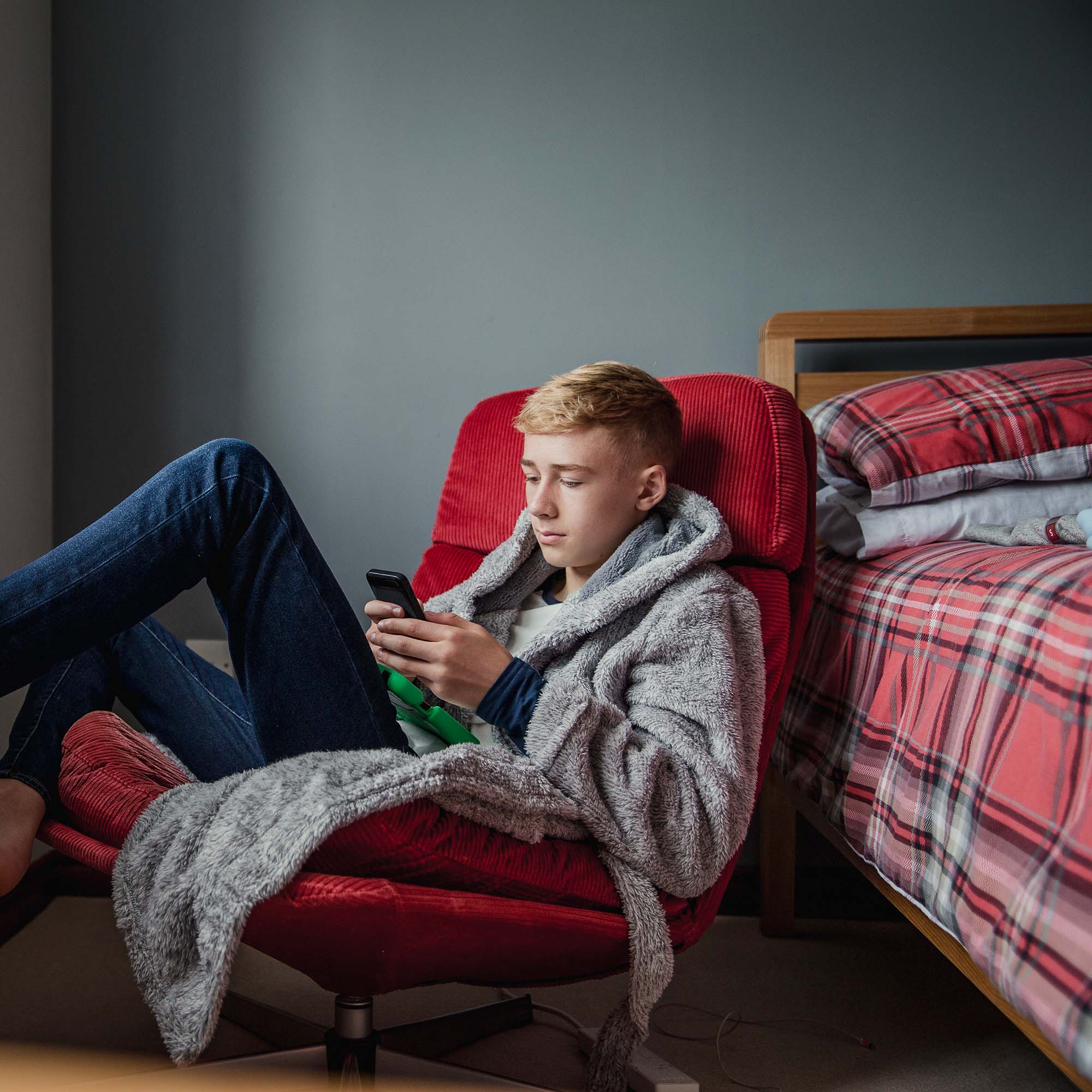 A teenage boy slouches in a chair next to his bed while he uses his cell phone.