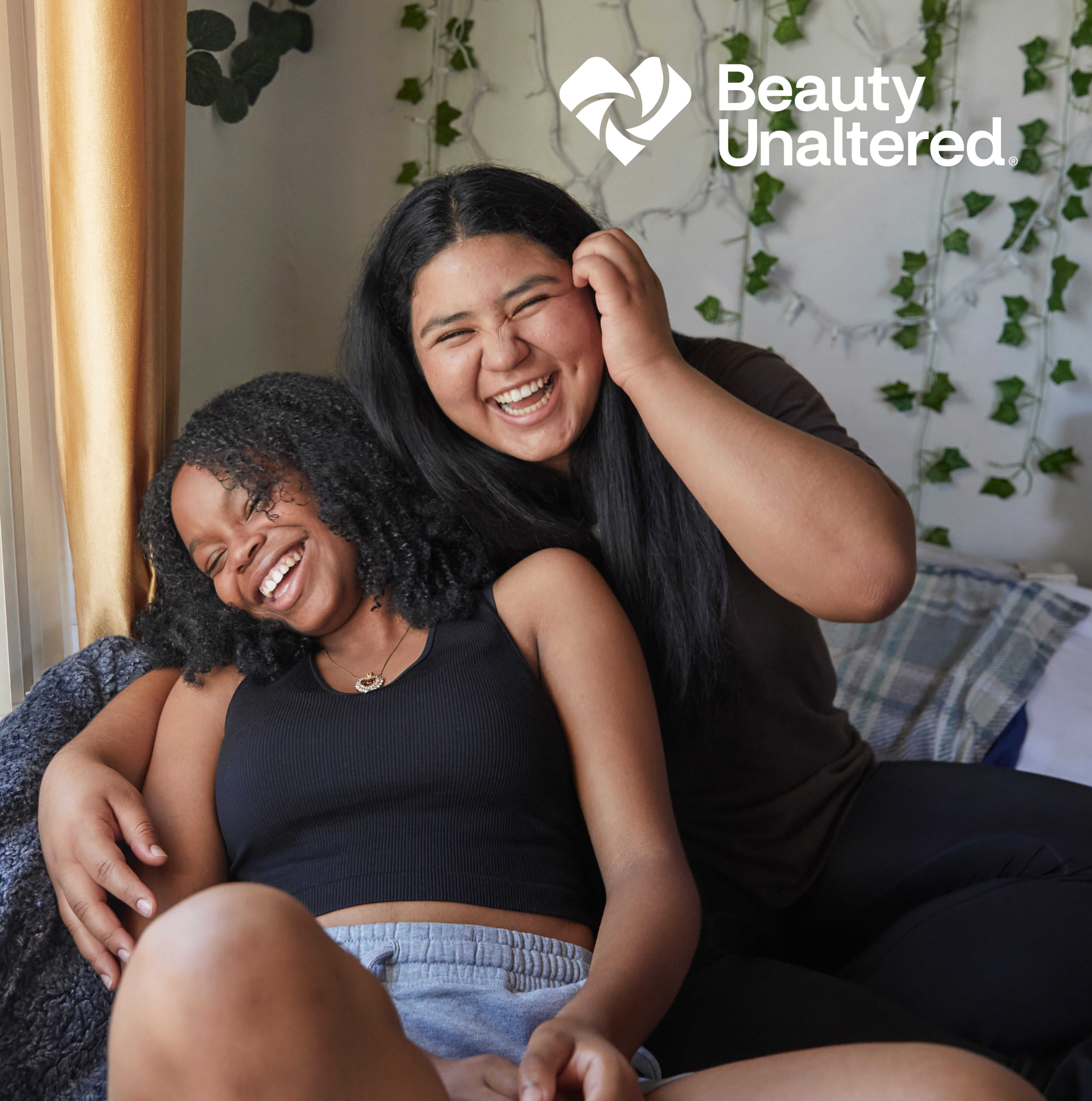 Two young naturally beautiful women of color hang out and laught together. The logo for beauty unaltered is shown.